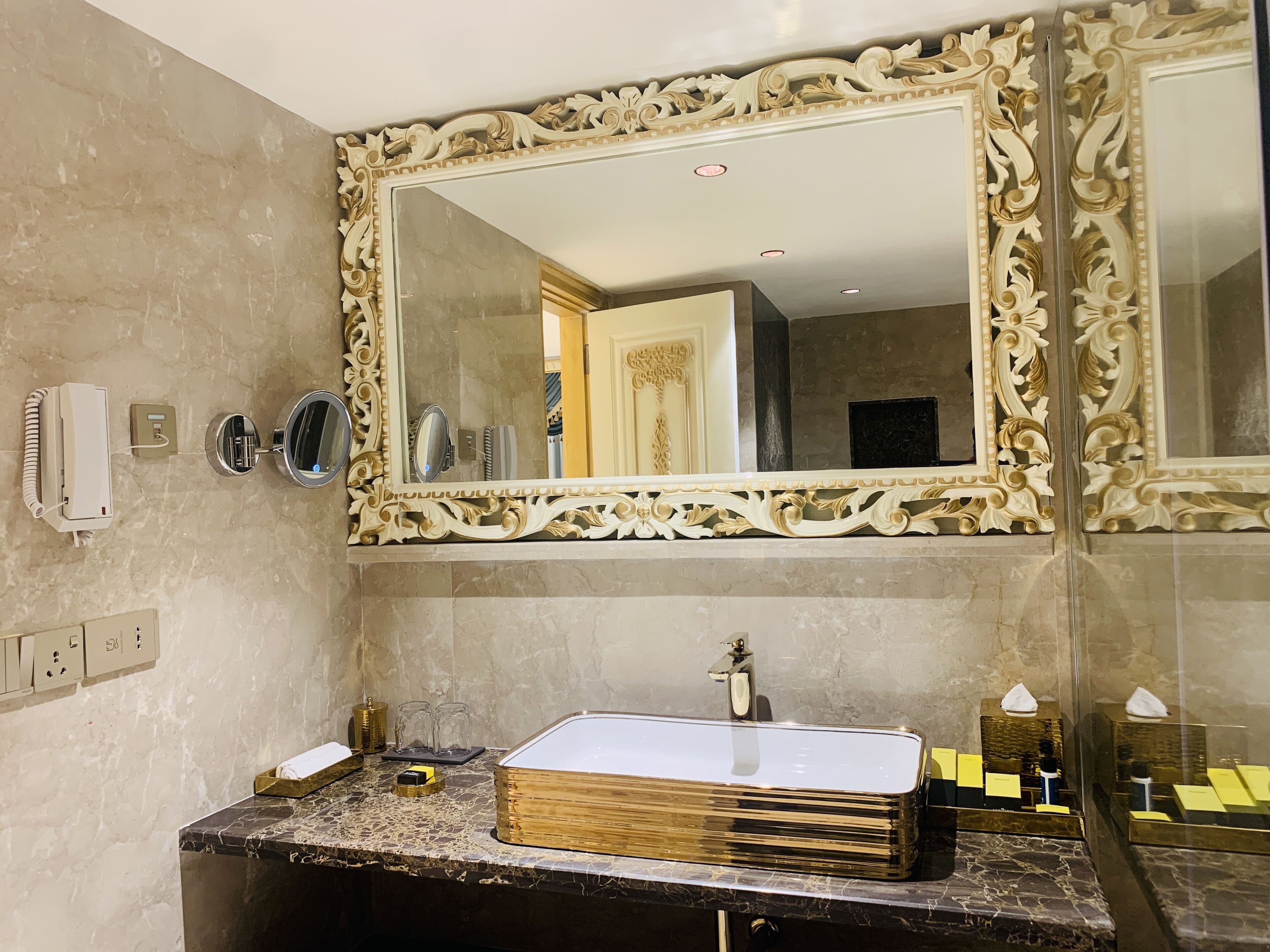 Ein Badezimmer mit einem großen, verzierten Spiegel, einer Marmorarbeitsplatte und einem Waschbecken mit goldener Oberfläche.