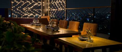 A dining area with a wooden table and chairs, a view of the cityscape at night, and decorative lights on the walls.