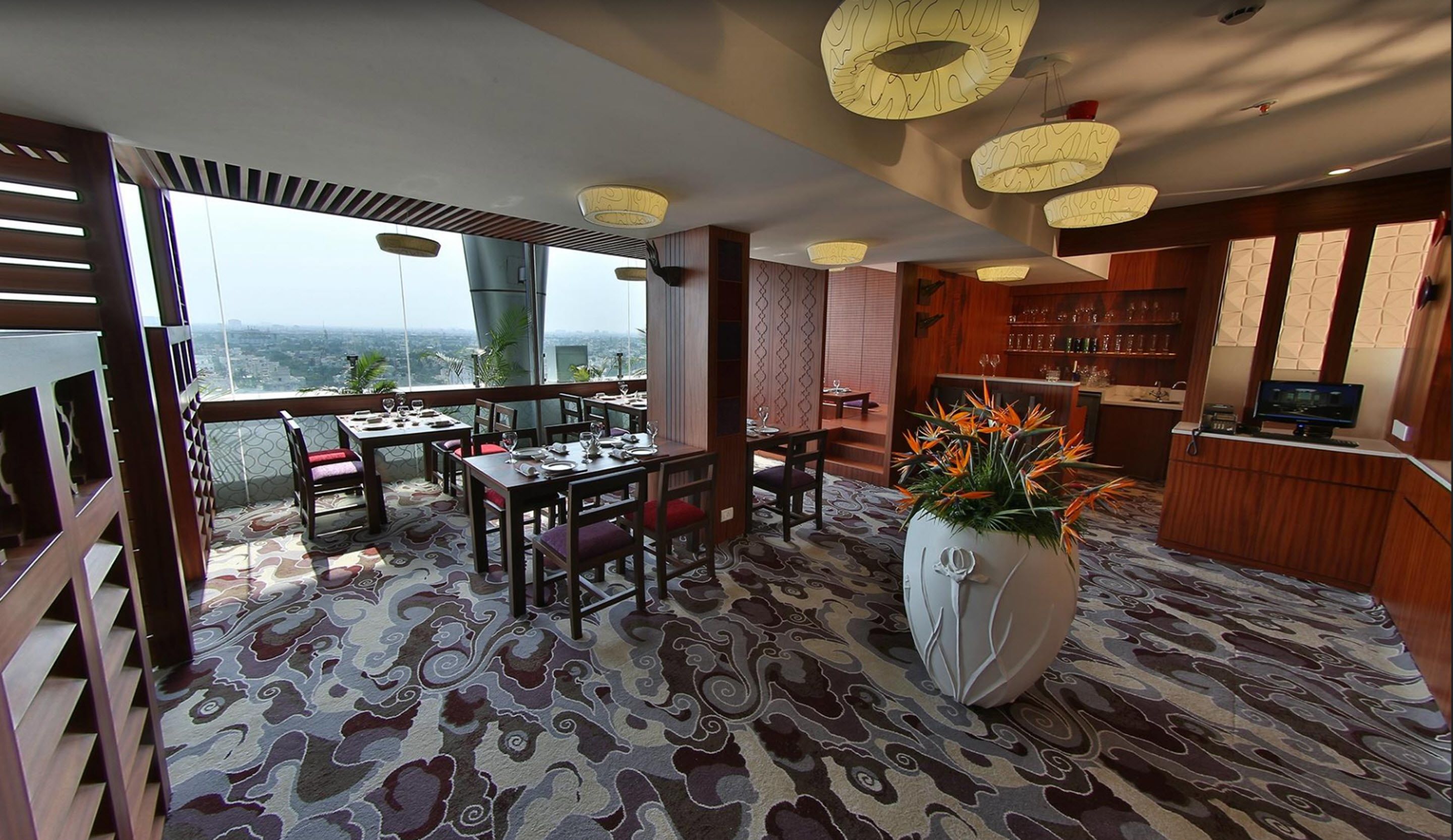 A dining area with a view, featuring a patterned carpet, wooden furniture, and a large vase with flowers.