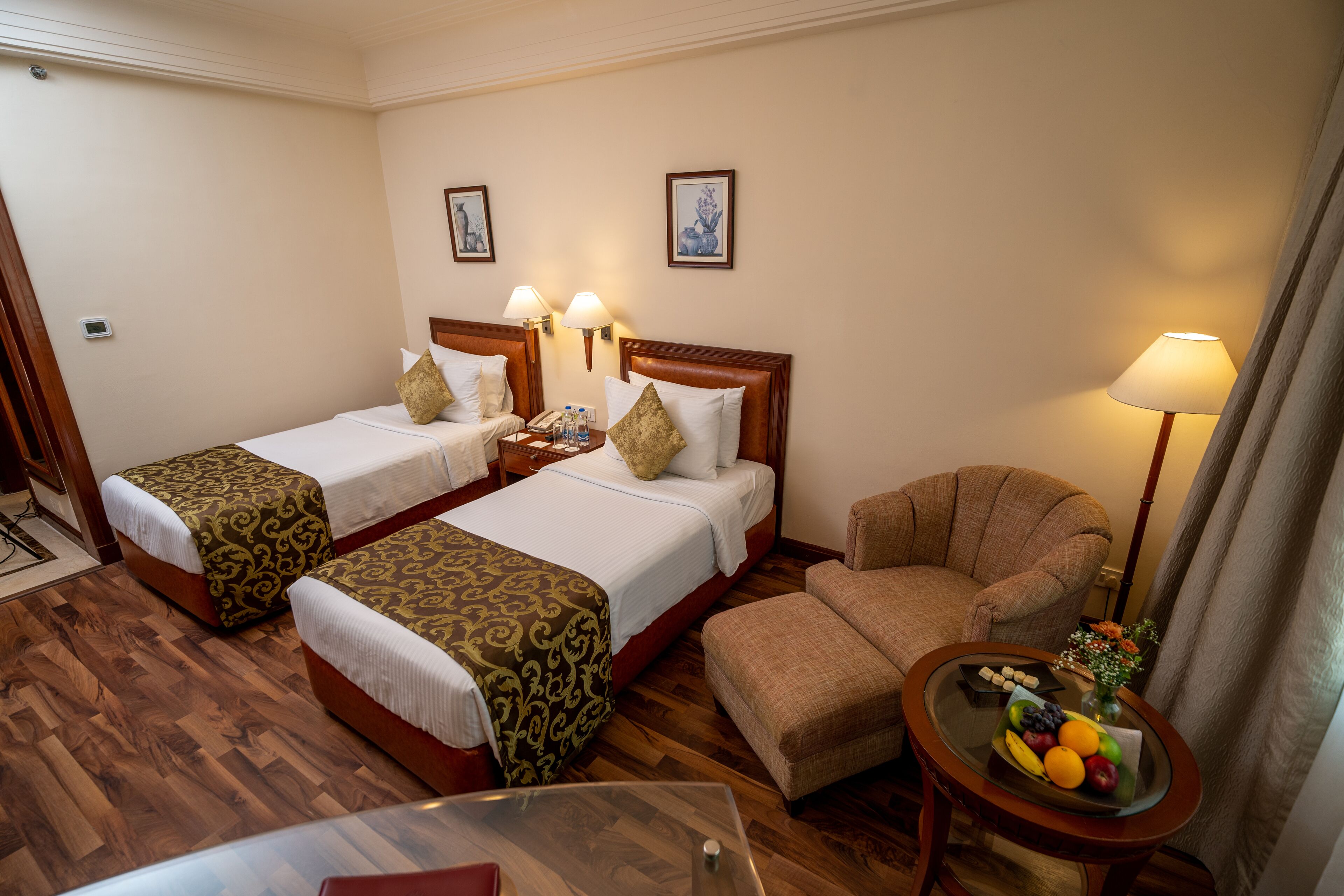 A hotel room with two beds, a sofa, a small table with fruit, and a lamp.