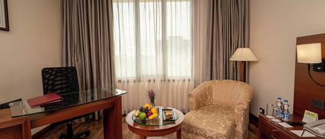 A hotel room with a wooden desk, a comfortable armchair, a small round table with fruit, and a lamp.