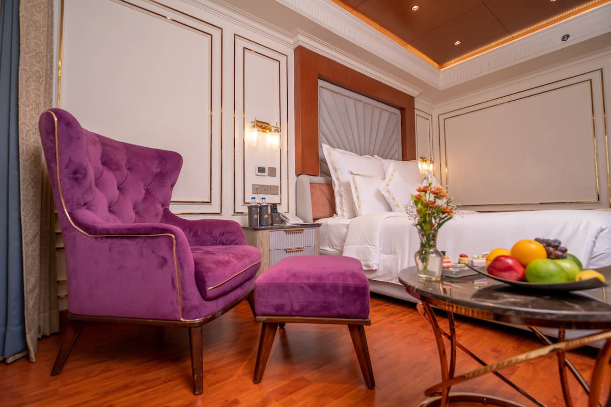 Habitación de hotel con una cama grande, un sillón púrpura, una mesita con fruta y una mesita de noche con lámpara.