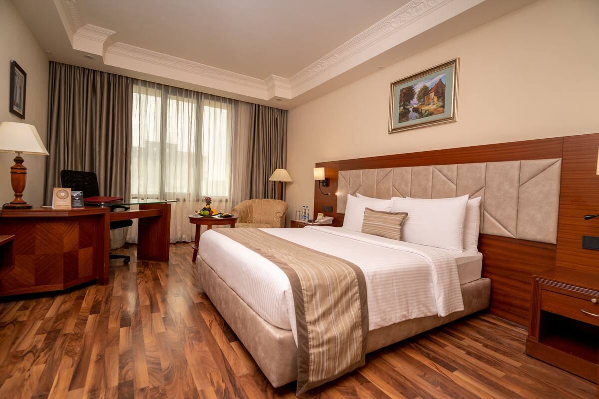 Habitación de hotel con una cama grande, suelo de madera, un escritorio con lámpara, una silla y un cuadro en la pared.