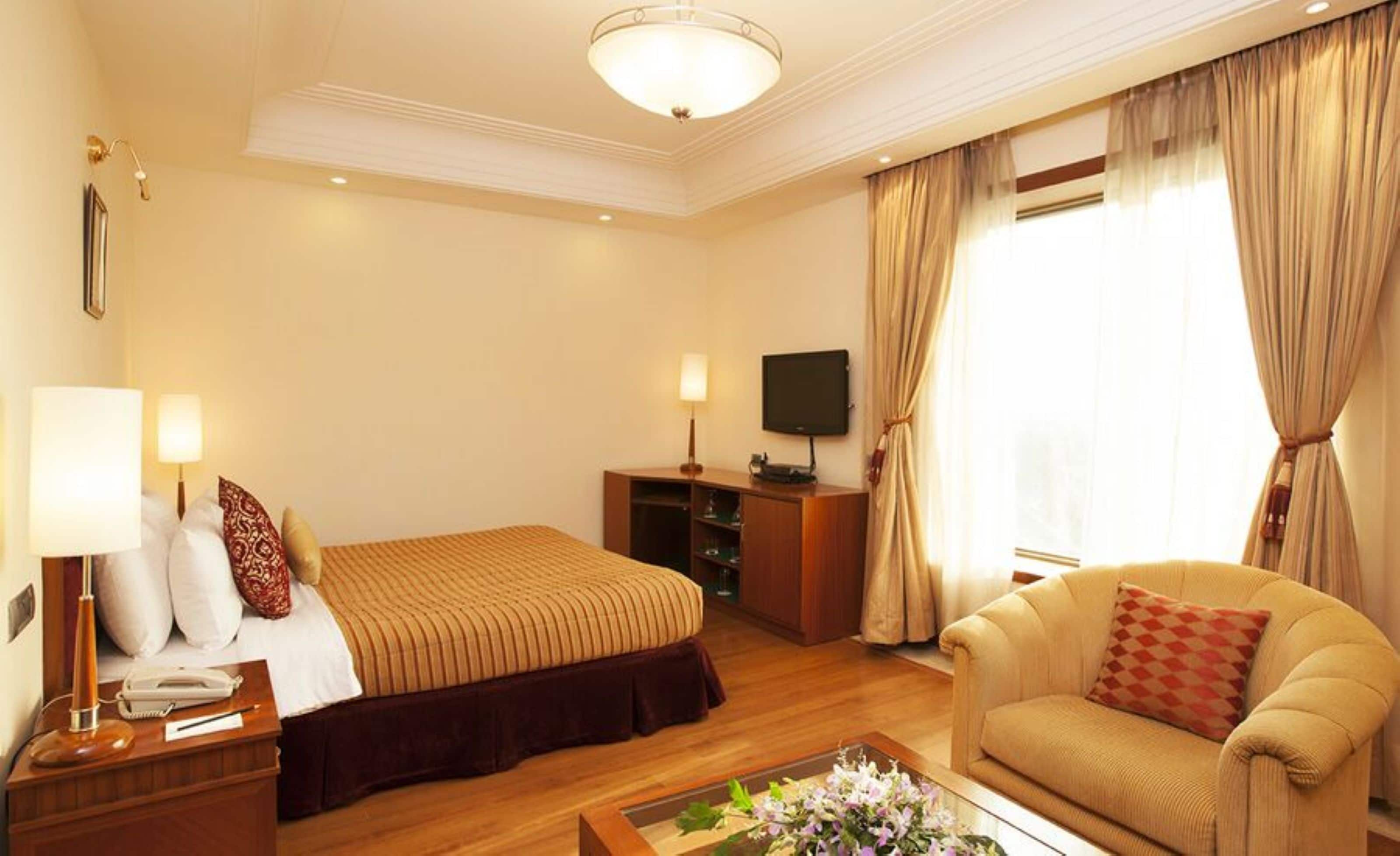 A hotel room with a bed, a sofa, a TV, a desk with a lamp, and a window with curtains.