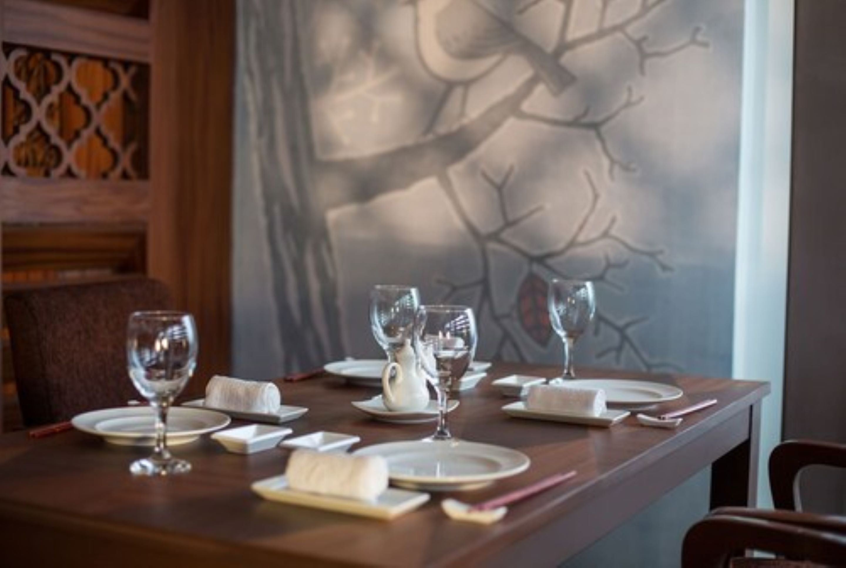 Una mesa de comedor servida con platos blancos, vasos y una tetera, con un mural de un árbol en la pared de fondo.