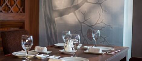 Una mesa de comedor servida con platos blancos, vasos y una tetera, con un mural de un árbol en la pared de fondo.