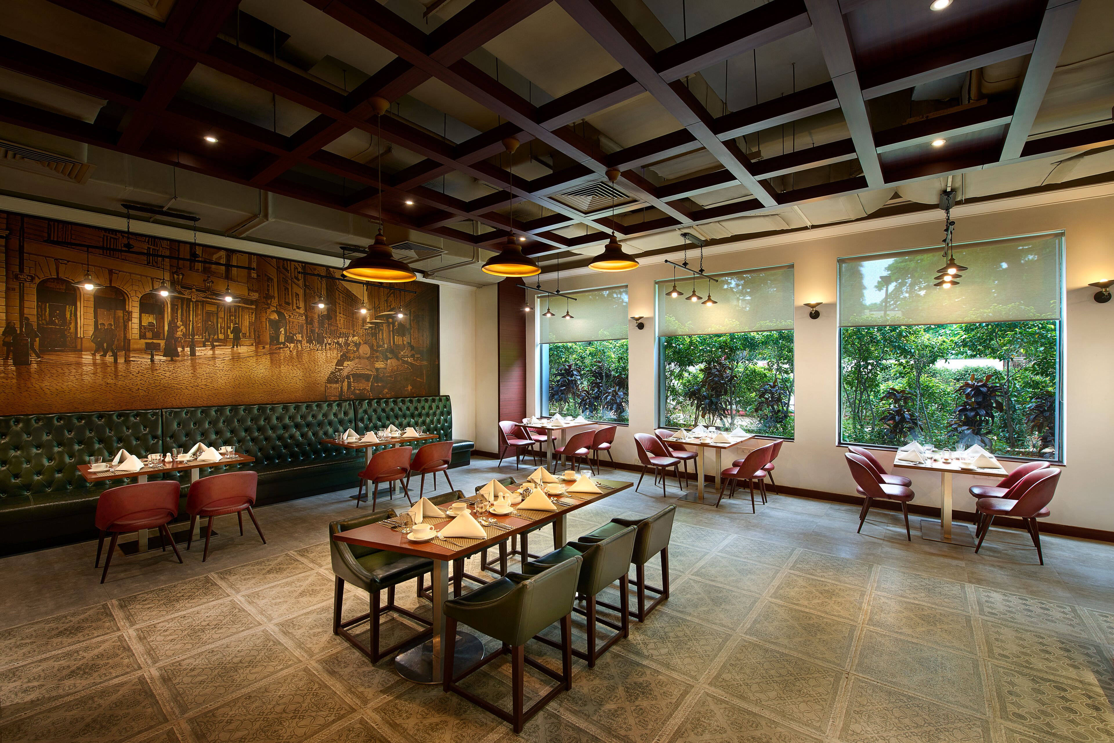 A restaurant with high ceilings, exposed beams, and large windows offering views of greenery. Tables are set with white tablecloths and red chairs.