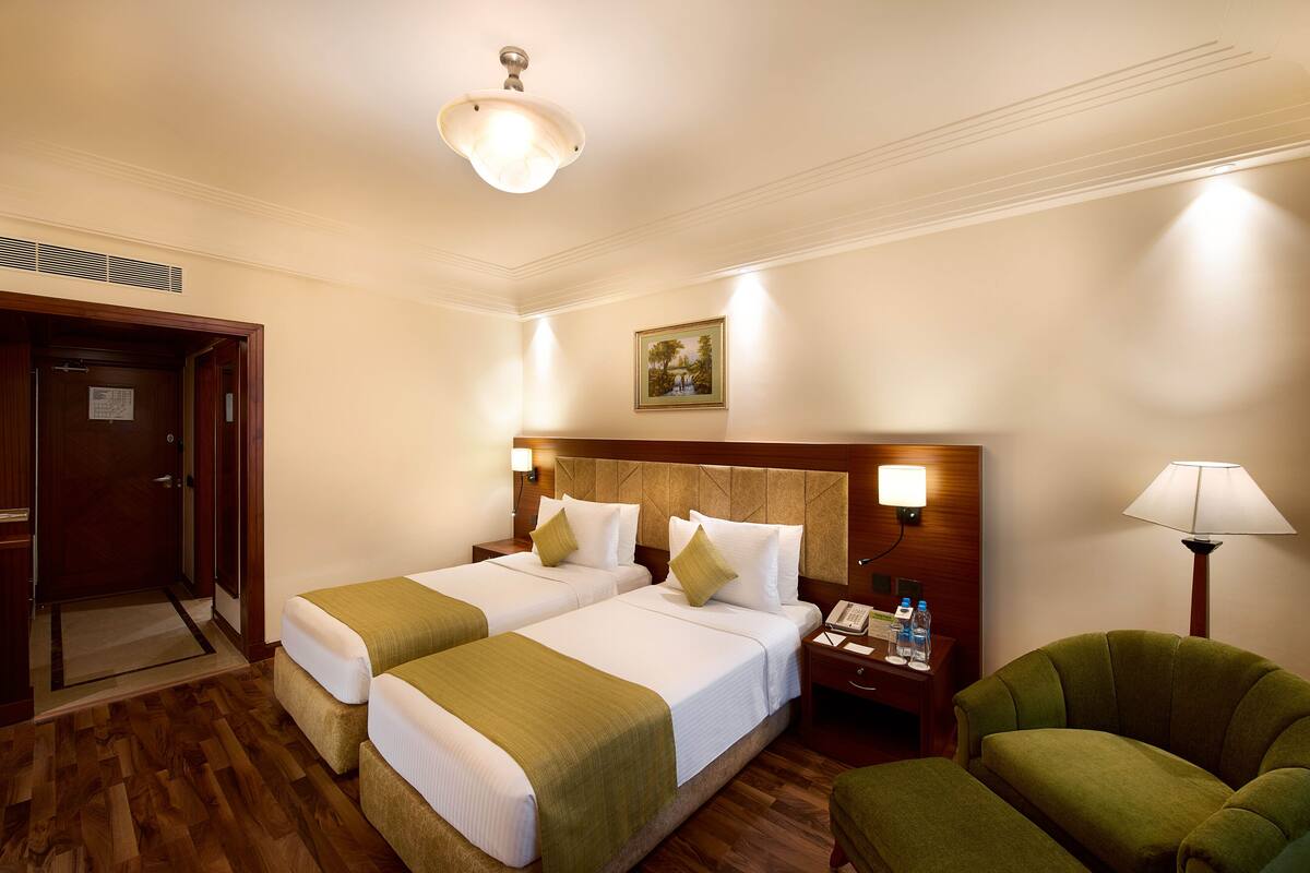Una habitación de hotel con dos camas, un cabecero de madera, una mesita de noche con lámpara, un sillón verde y una puerta.