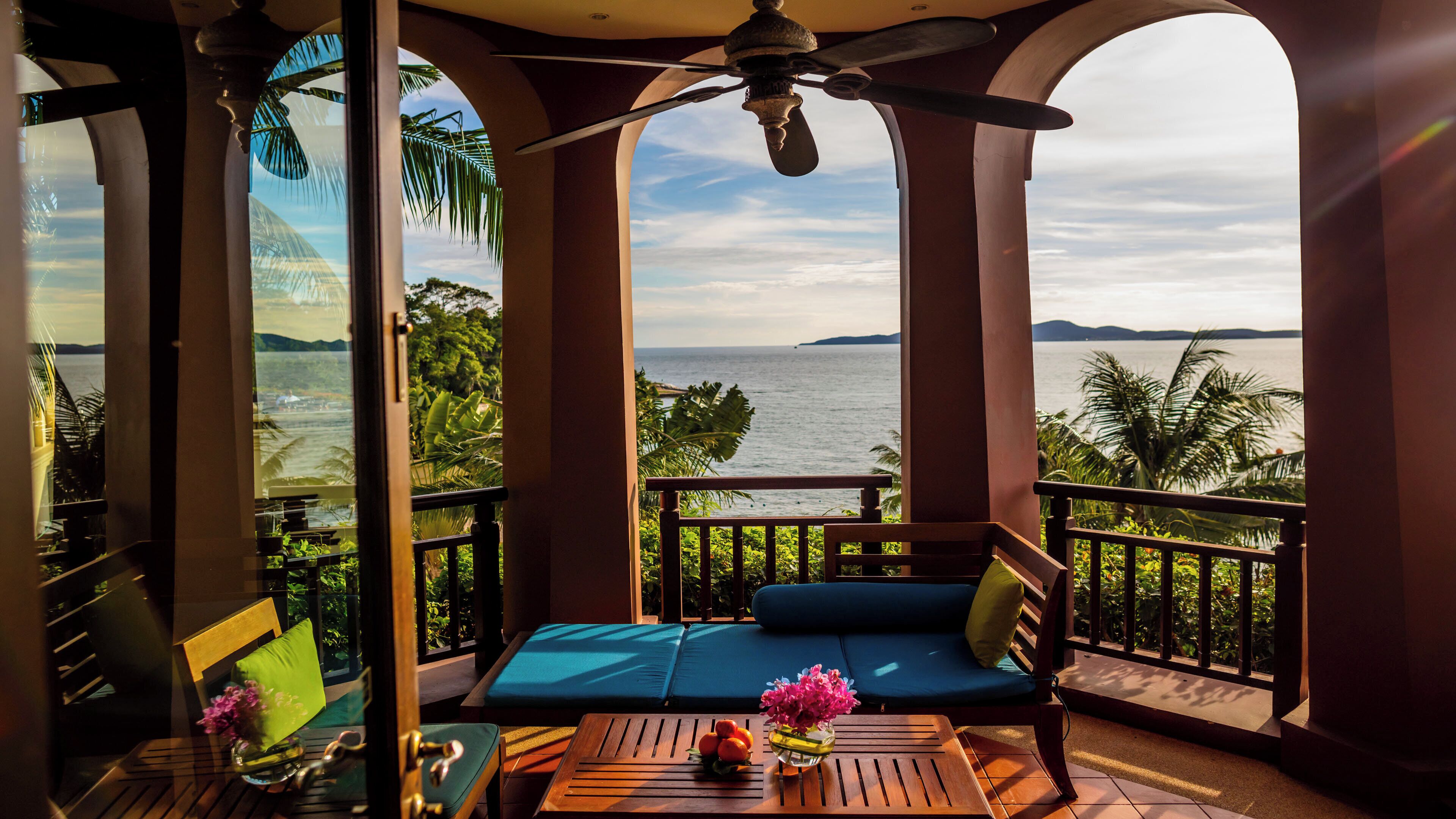 A balcony with a view of the sea, a ceiling fan, and a table with a vase of flowers.