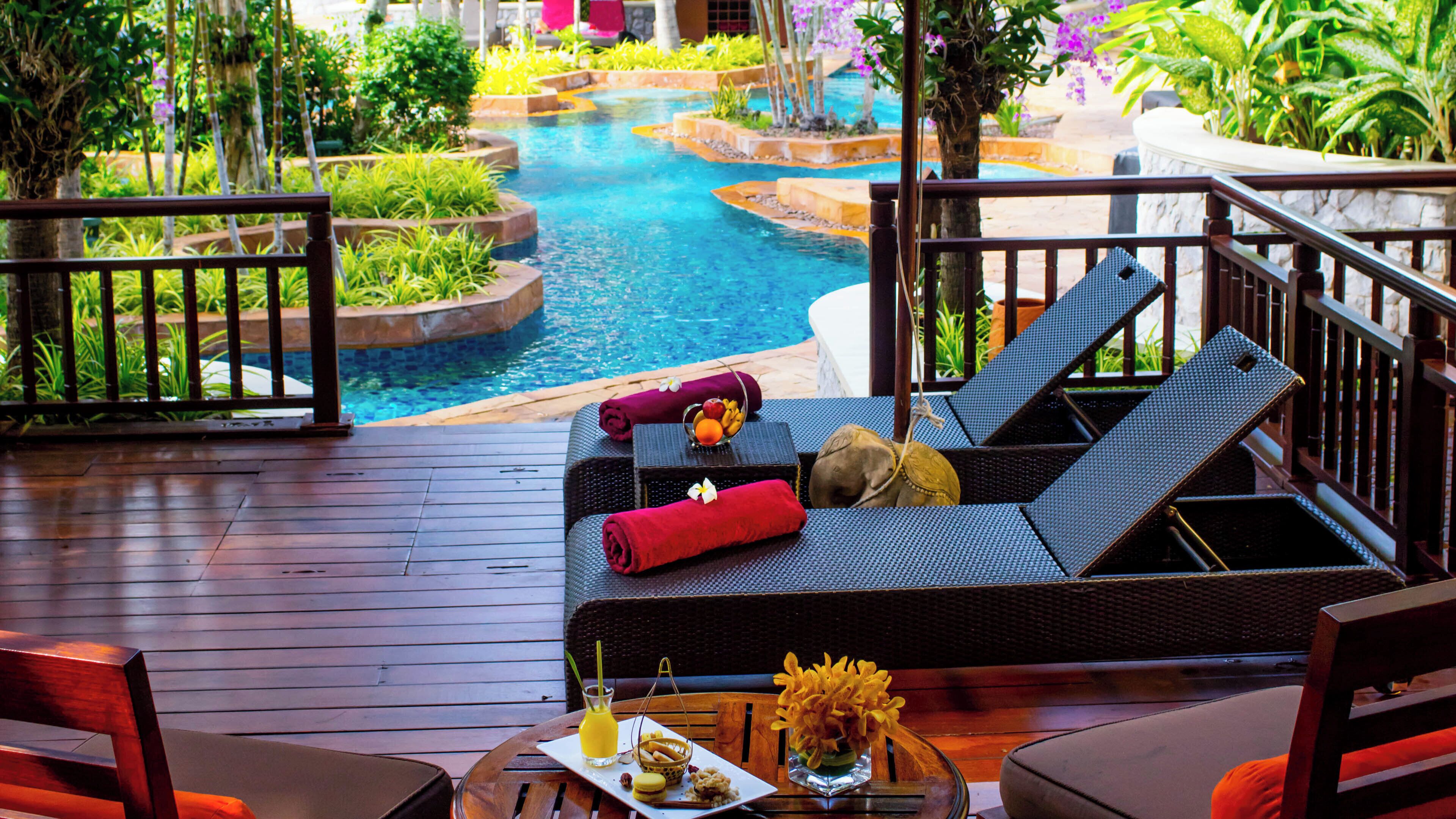 A poolside area with lounge chairs, a table with a drink and snacks, and a view of a water feature.