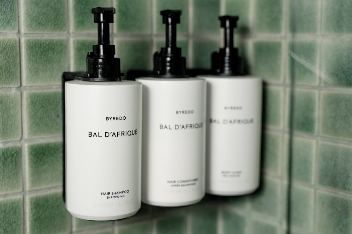 Three bottles of BYREDO BAL D'AFRIQUE hair shampoo against a green tiled wall.