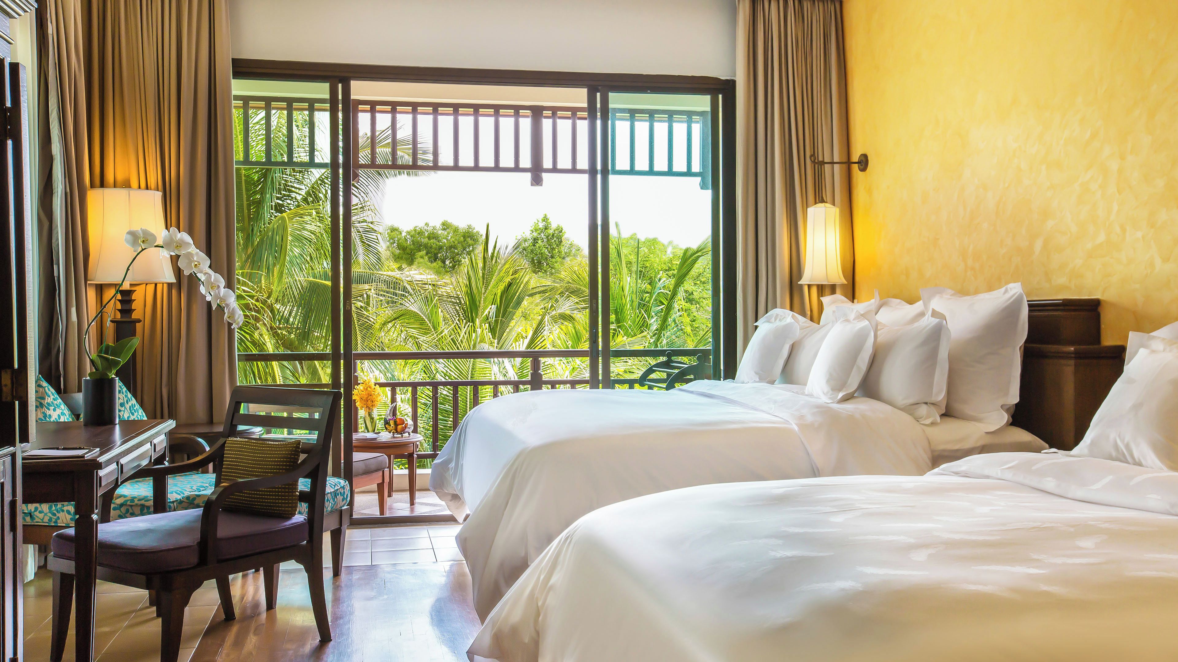 A hotel room with two beds, a desk with a chair, a small table with a vase, and a view of greenery through large windows.