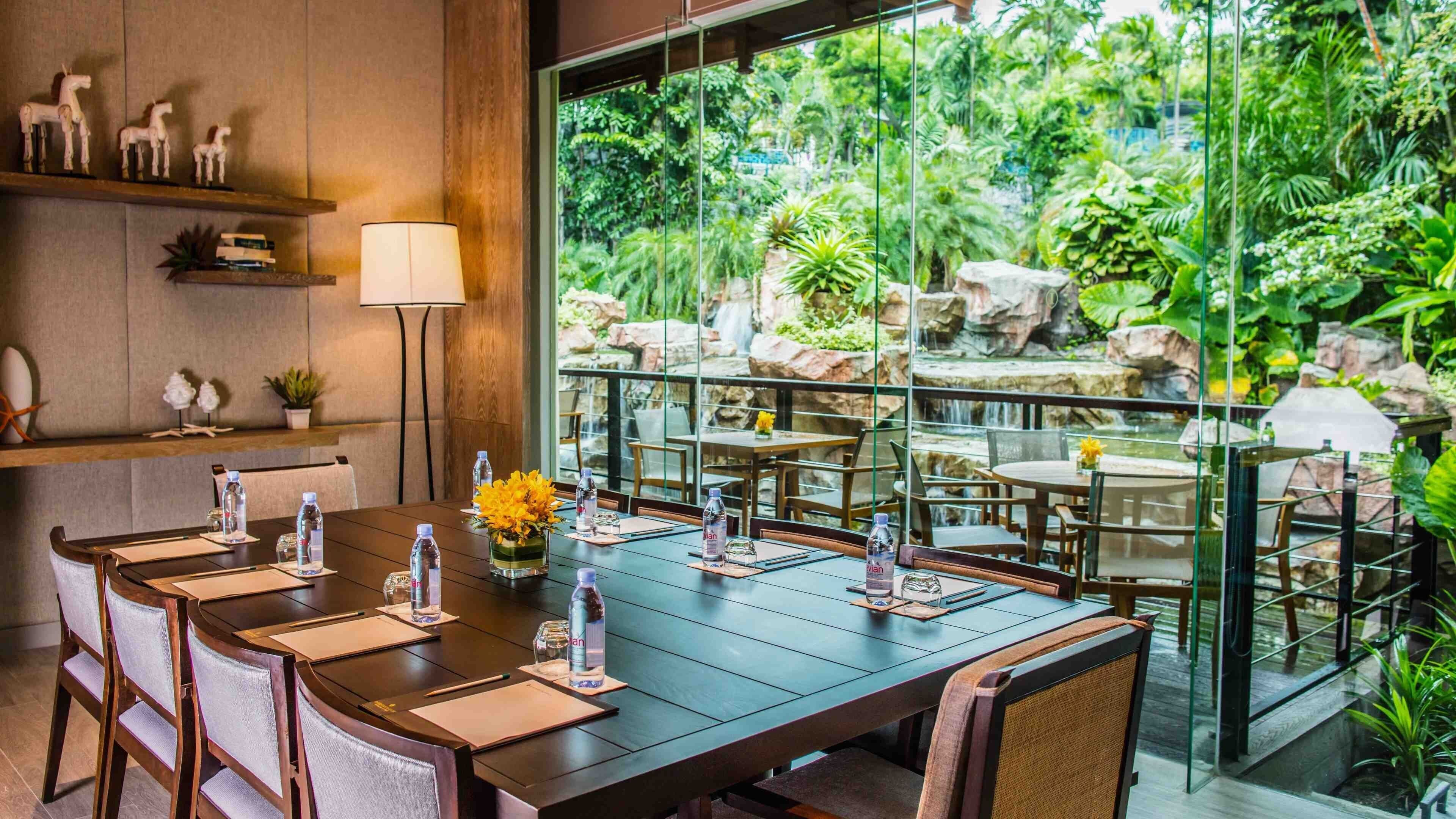 Une salle de conférence dotée d’une grande table, de chaises et offrant une vue sur un jardin luxuriant avec une cascade.