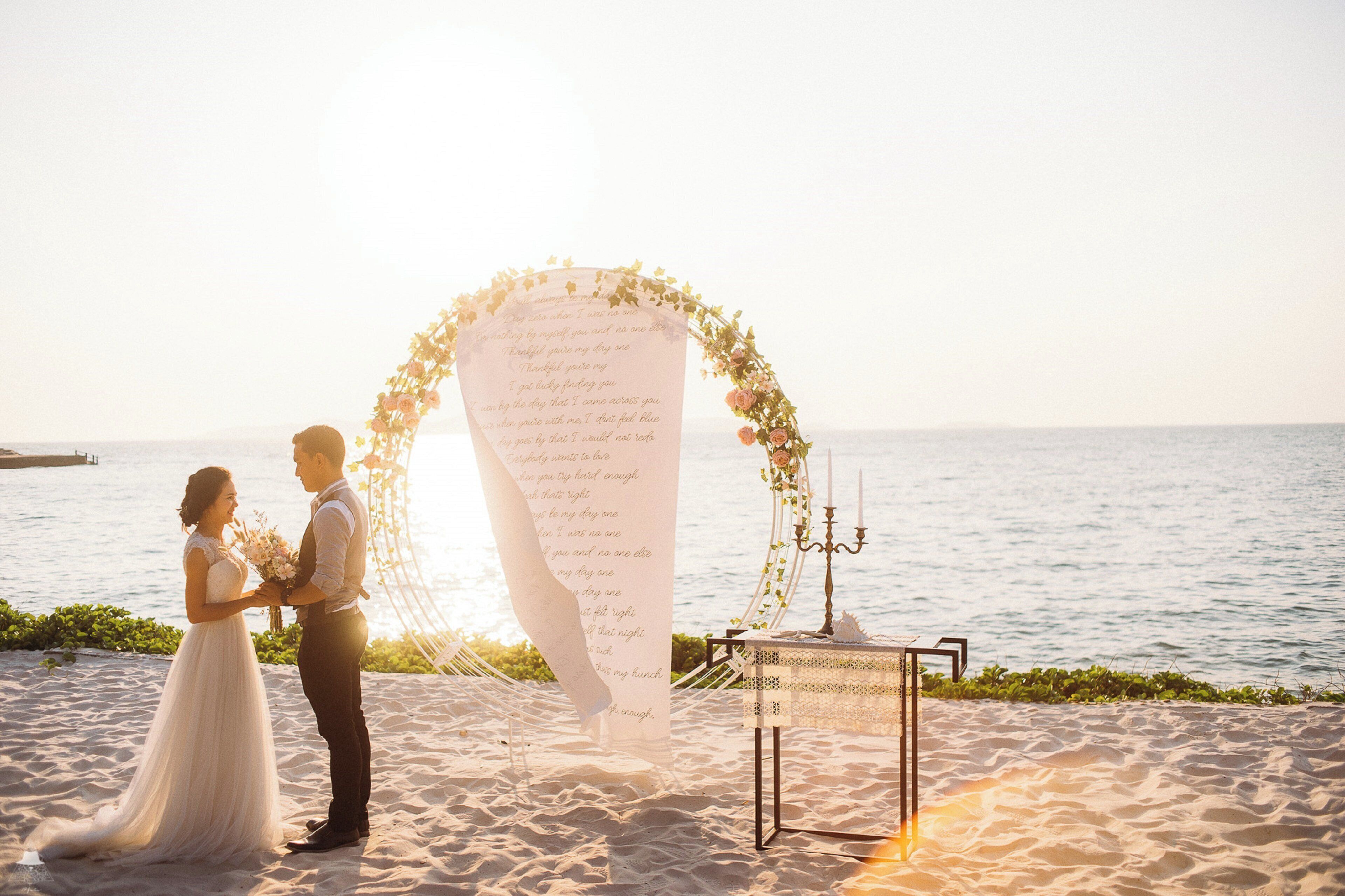 A couple standing in front of a beach wedding arch with a floral arrangement and a table with candles.