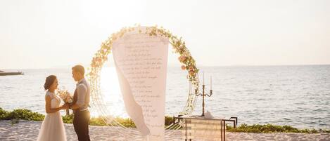 A couple standing in front of a beach wedding arch with a floral arrangement and a table with candles.