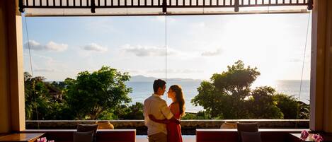 Una pareja abrazándose en una habitación con un amplio ventanal que ofrece una vista panorámica del mar y la vegetación.
