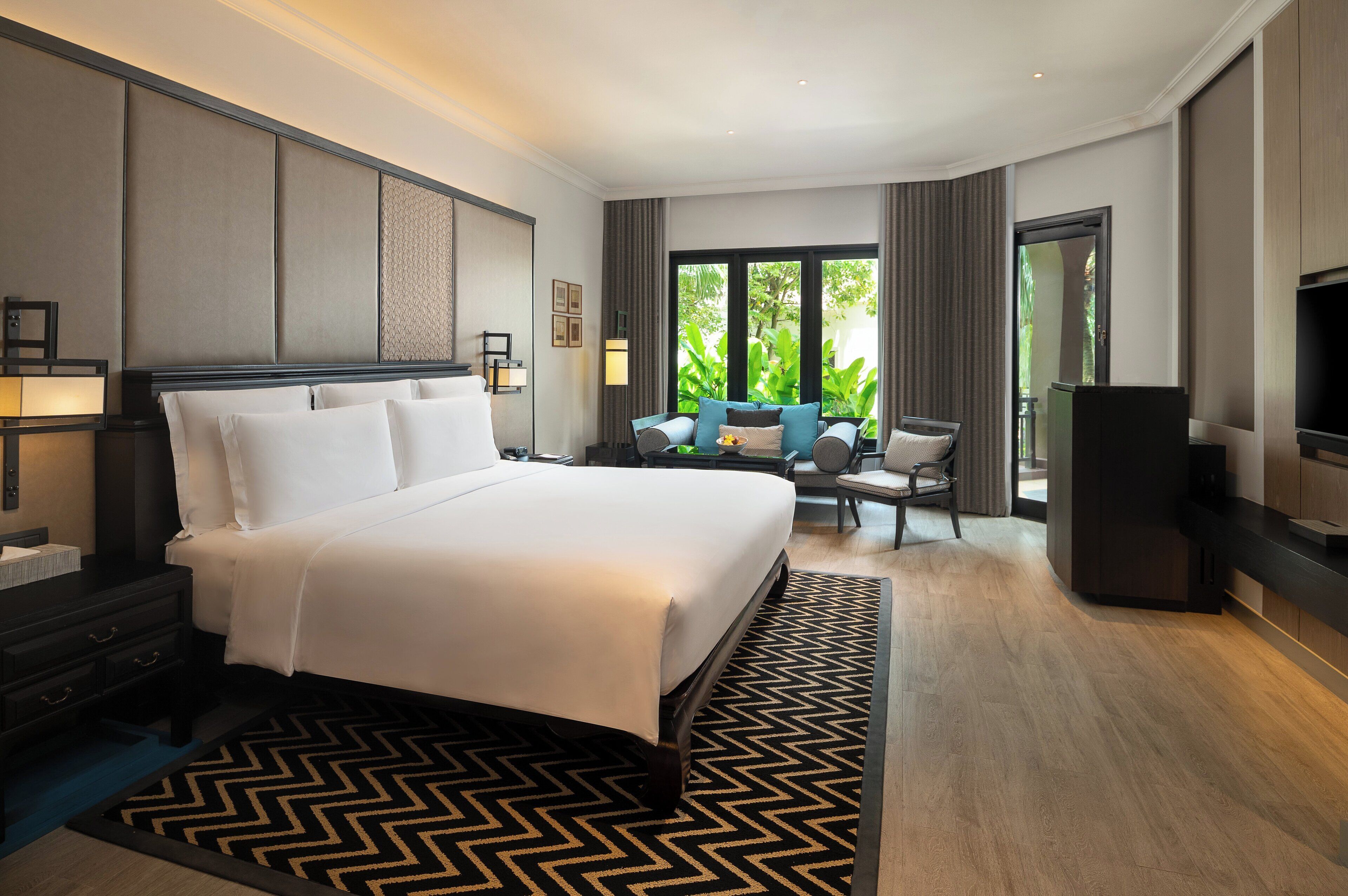A hotel room with a large bed, a desk, a chair, a TV, and a view of greenery through the windows.