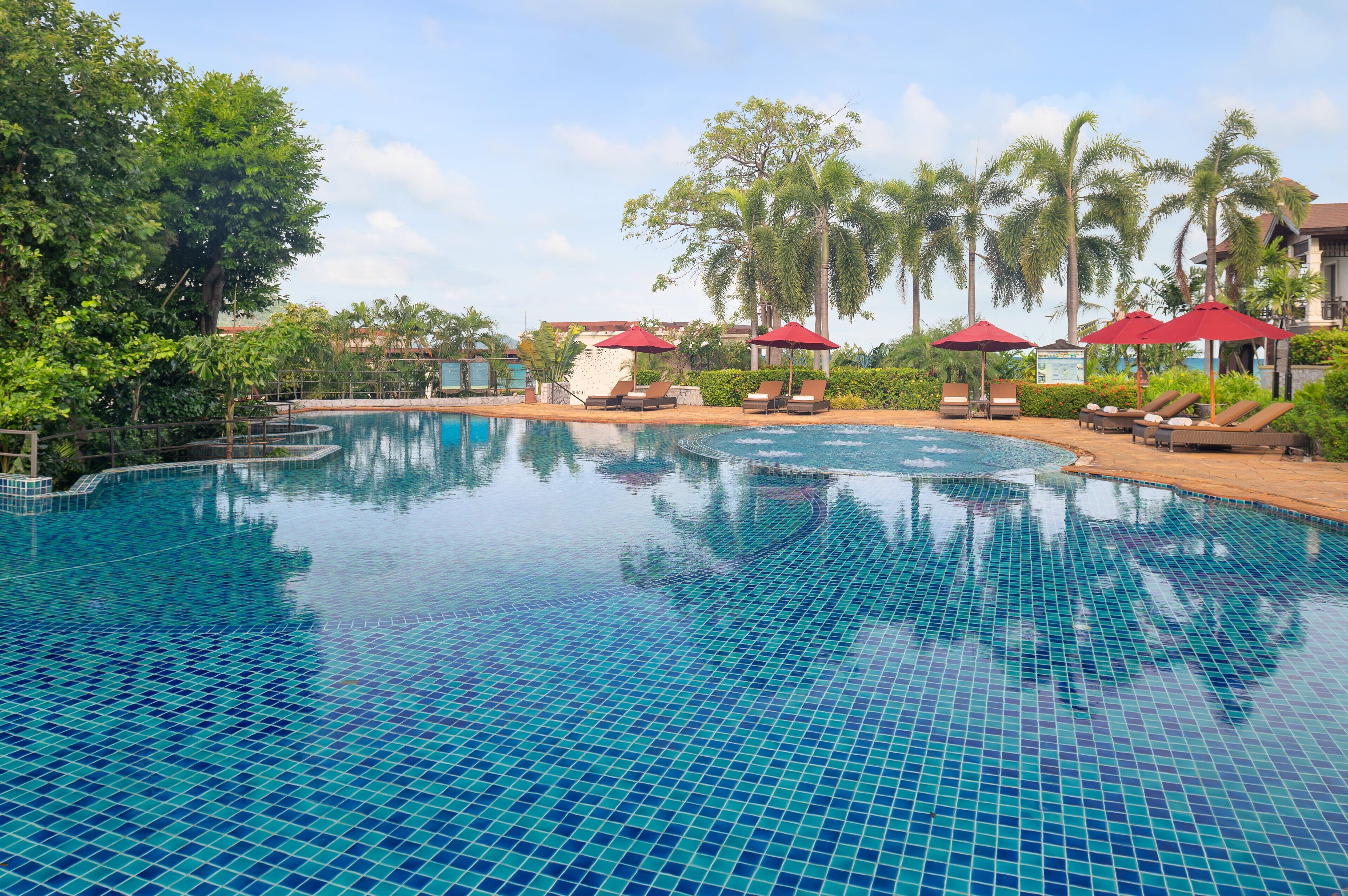 A swimming pool with tiled blue surface, surrounded by lounge chairs and red umbrellas, with palm trees and a building in the background.