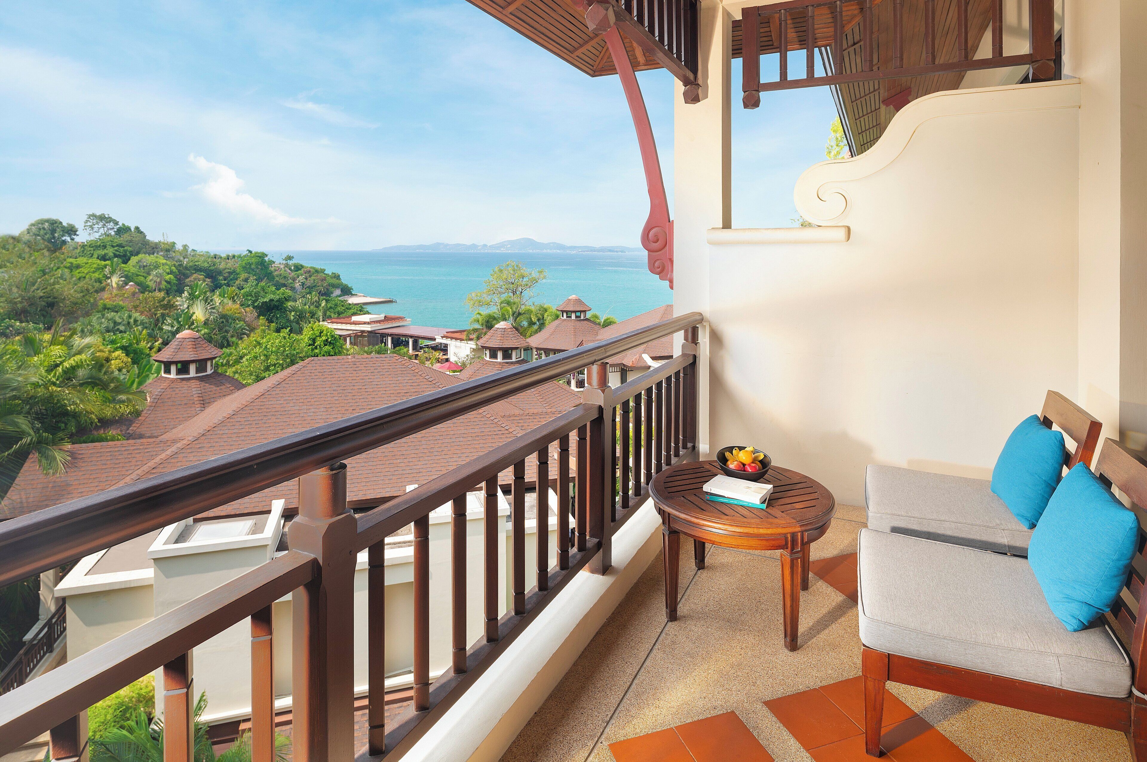 A balcony with a view of the sea, a round wooden table with a bowl of fruit, and a sofa with cushions.