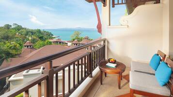 A balcony with a view of the sea, a round wooden table with a bowl of fruit, and a sofa with cushions.