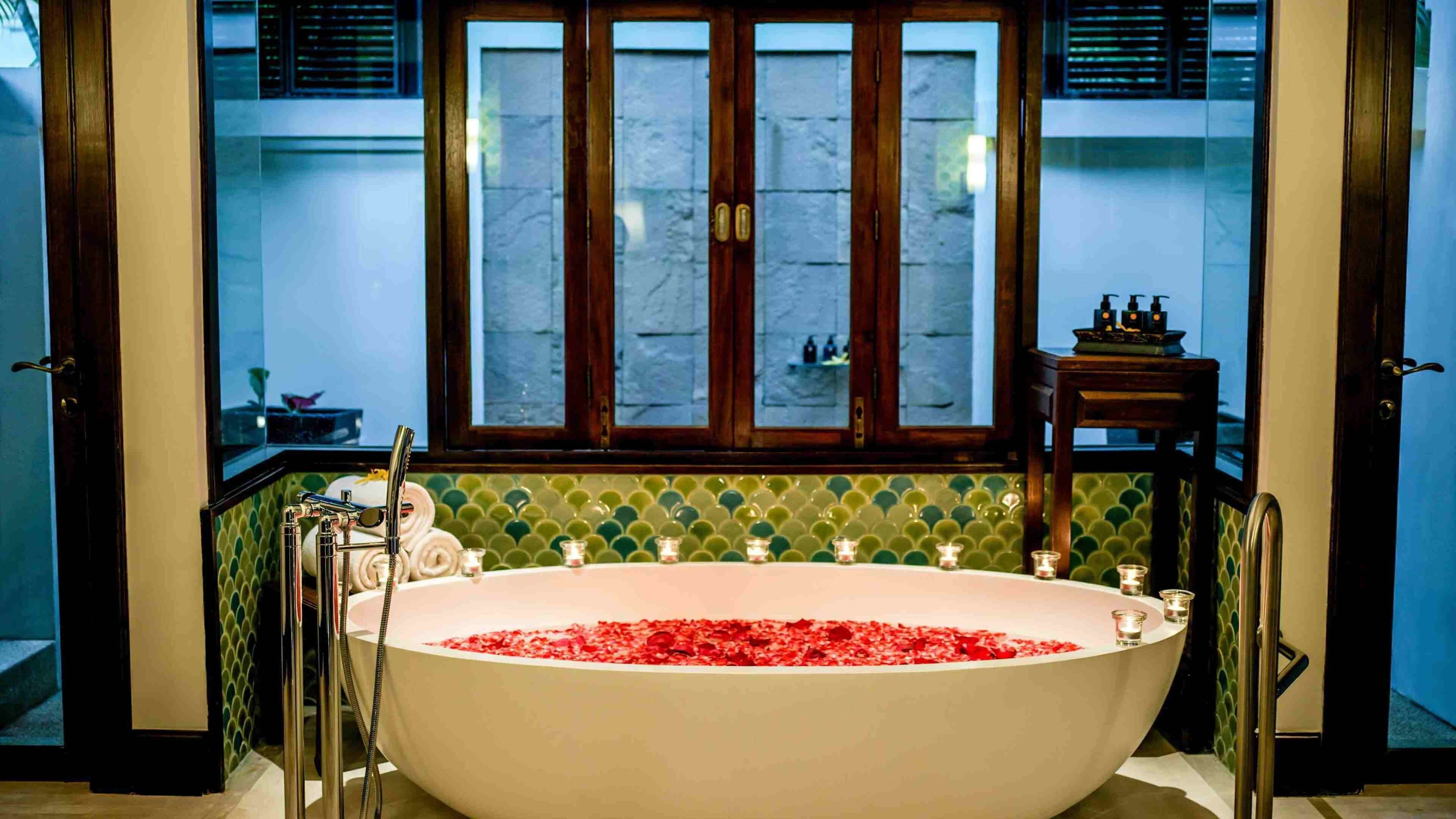 A large freestanding bathtub filled with rose petals, a window with a view of the street, and a small table with toiletries.