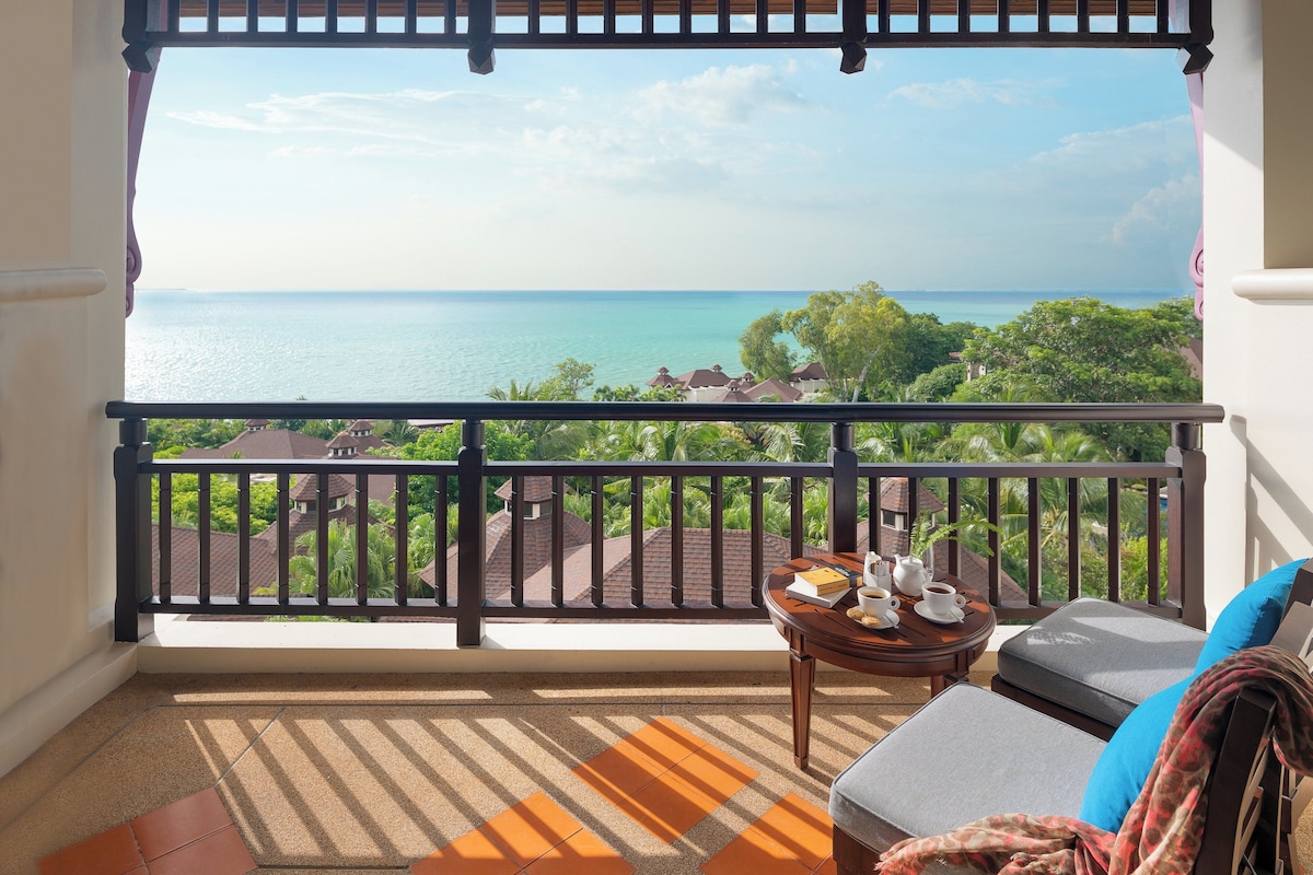 A balcony with a view of the sea, a table with a cup and saucer, and a sofa with cushions.