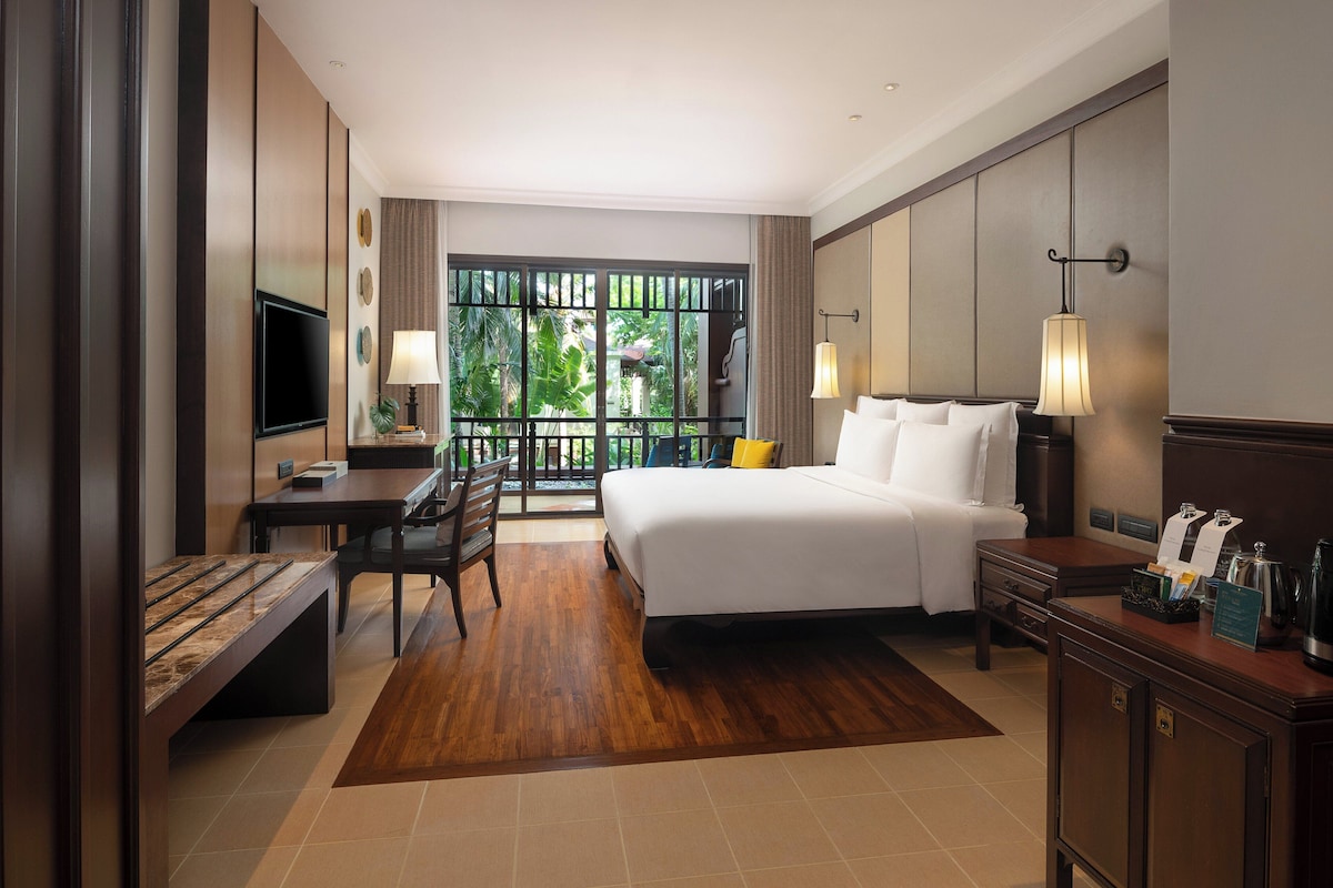 A hotel room with a large bed, a desk with a TV, a bench, and a view of greenery through the window.