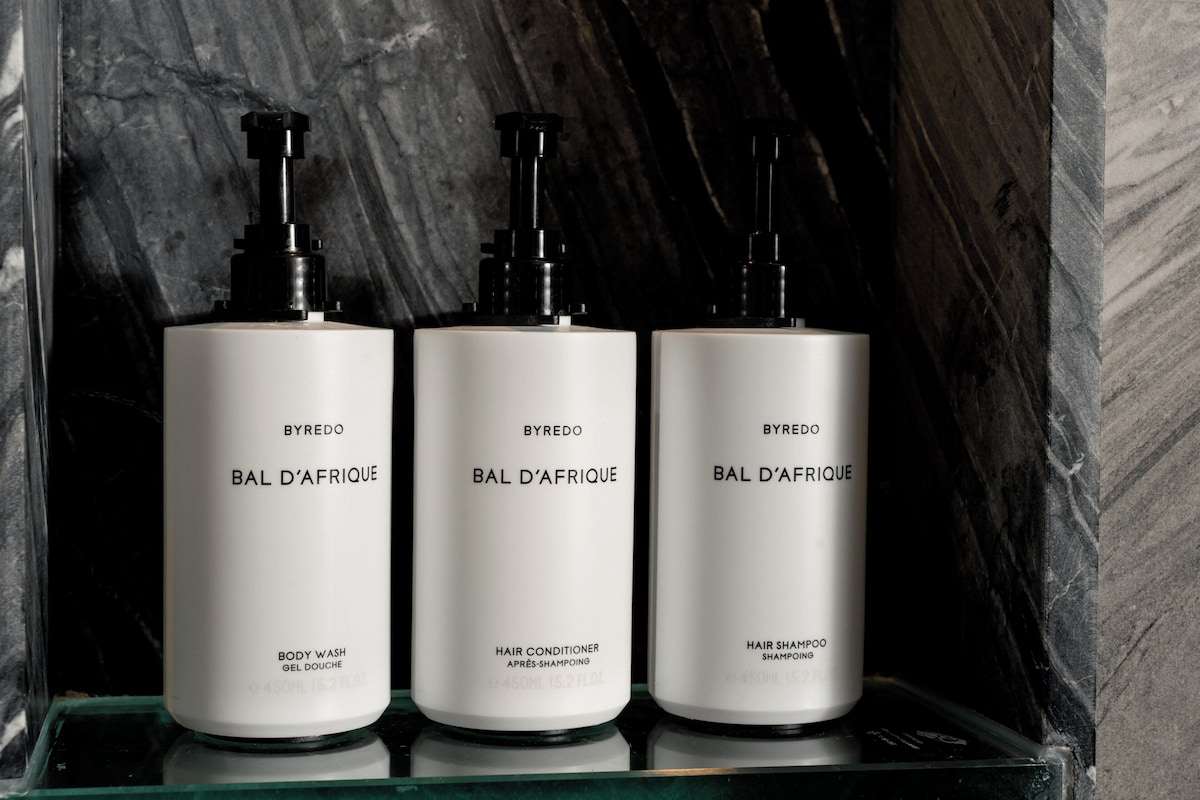 Three white bottles of BYREDO Bal D'Afrique body wash, hair conditioner, and shampoo on a glass shelf against a marble wall.