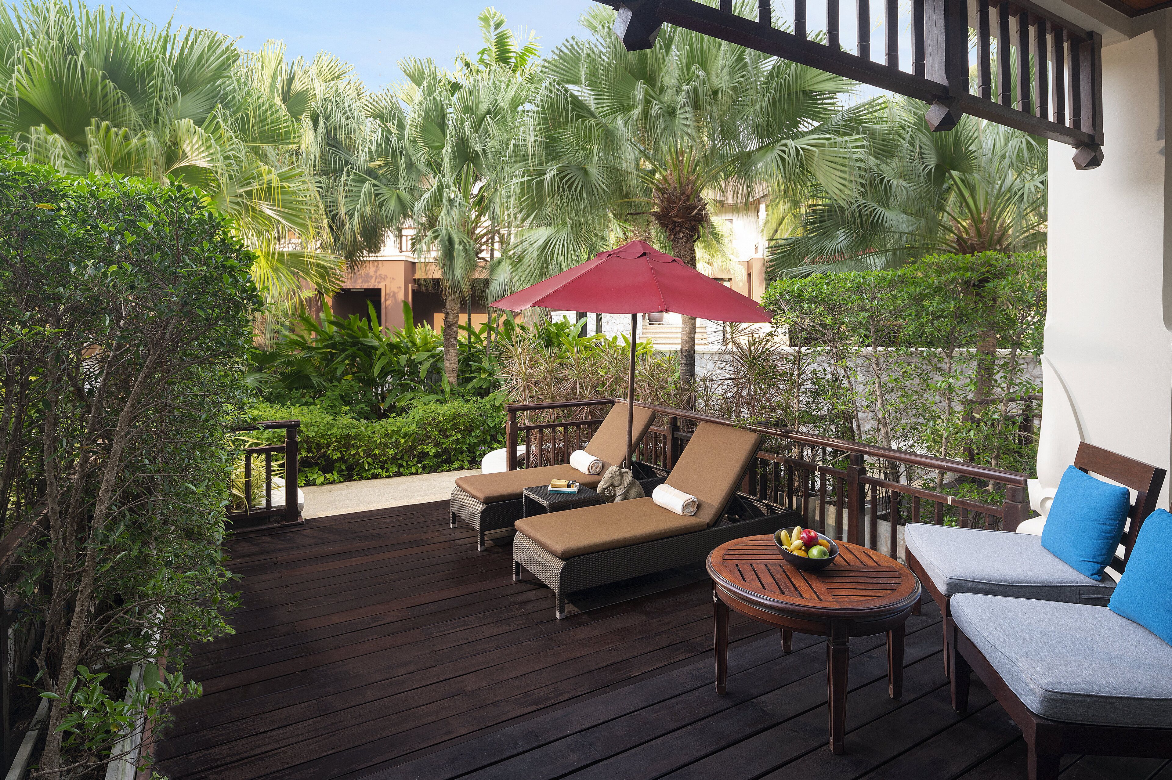 Een houten terras met twee lounge stoelen, een bijzettafeltje met fruit, en een rode parasol.