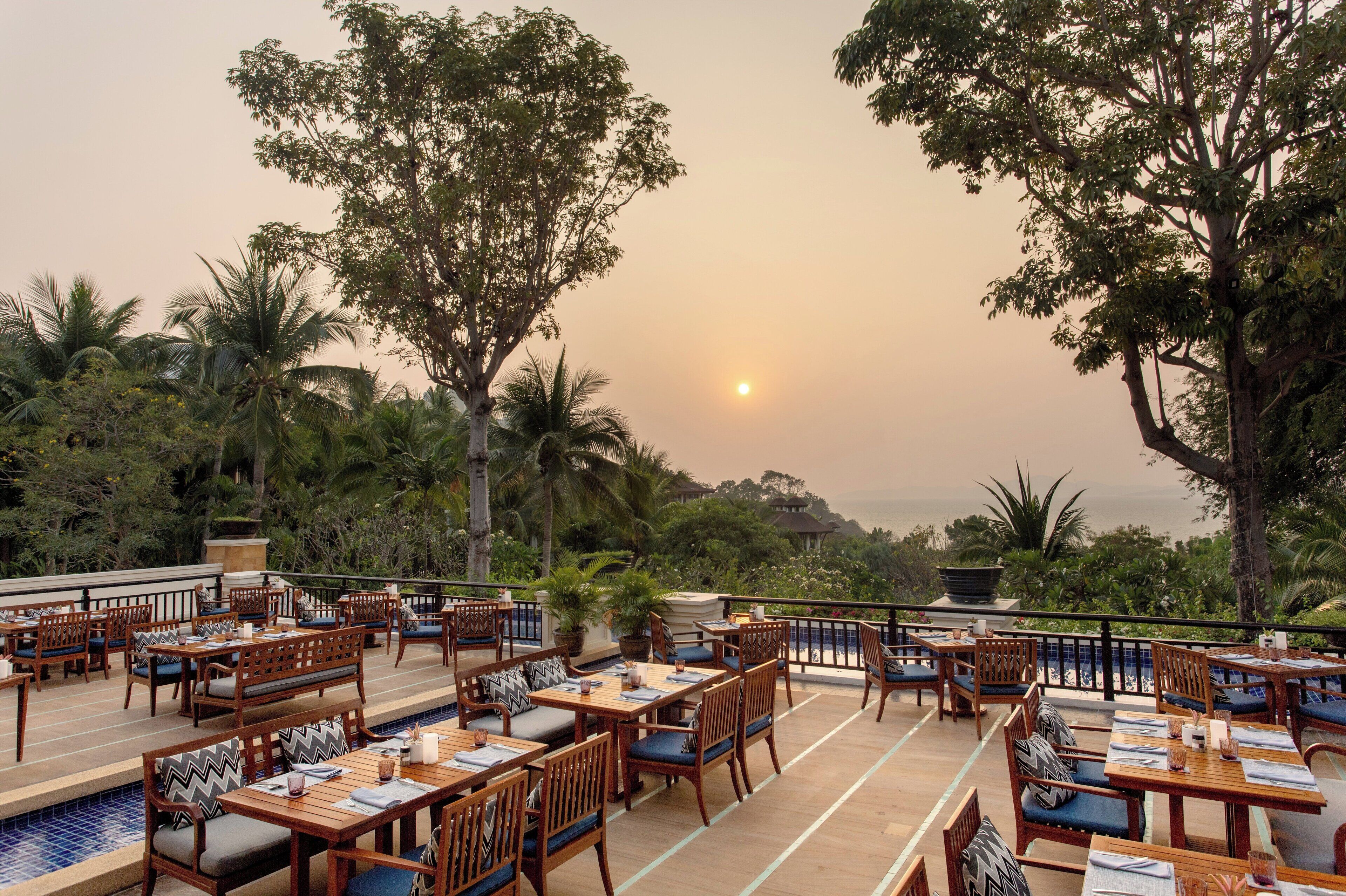 An outdoor dining area with wooden tables and chairs, surrounded by lush greenery and overlooking a scenic view.