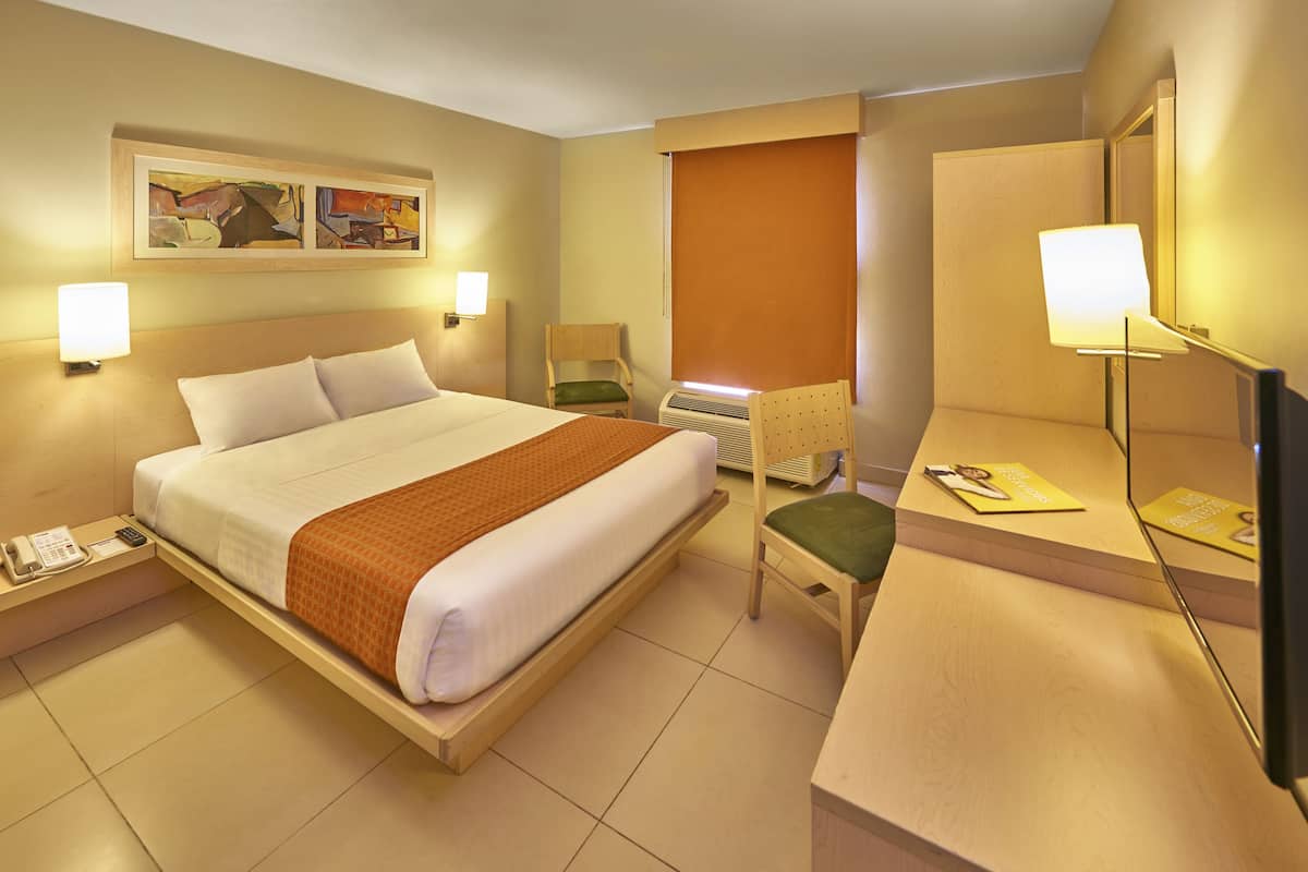 Ein Hotelzimmer mit Bett, Nachttisch, Lampe, Stuhl und Schreibtisch mit Spiegel.