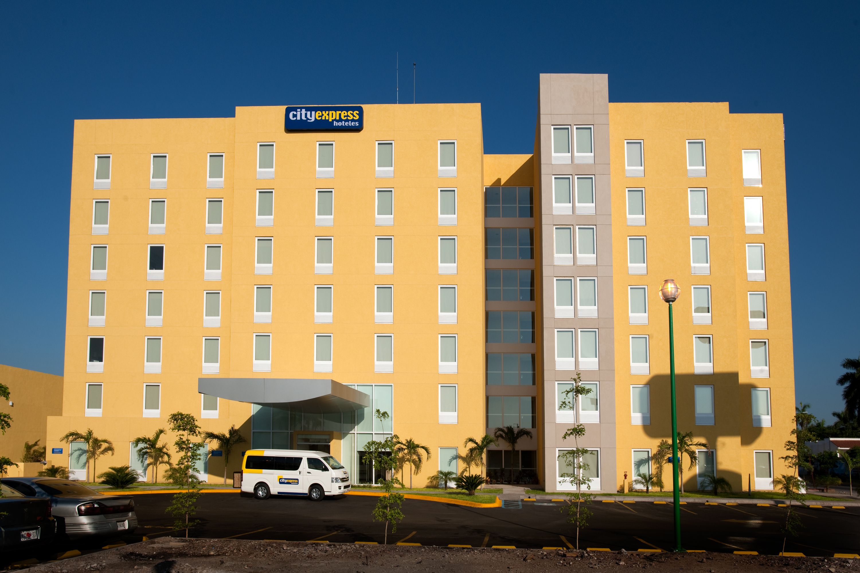 Un hotel de varios pisos con fachada amarilla, un letrero de 'CityExpress Hotels', un estacionamiento y palmeras.