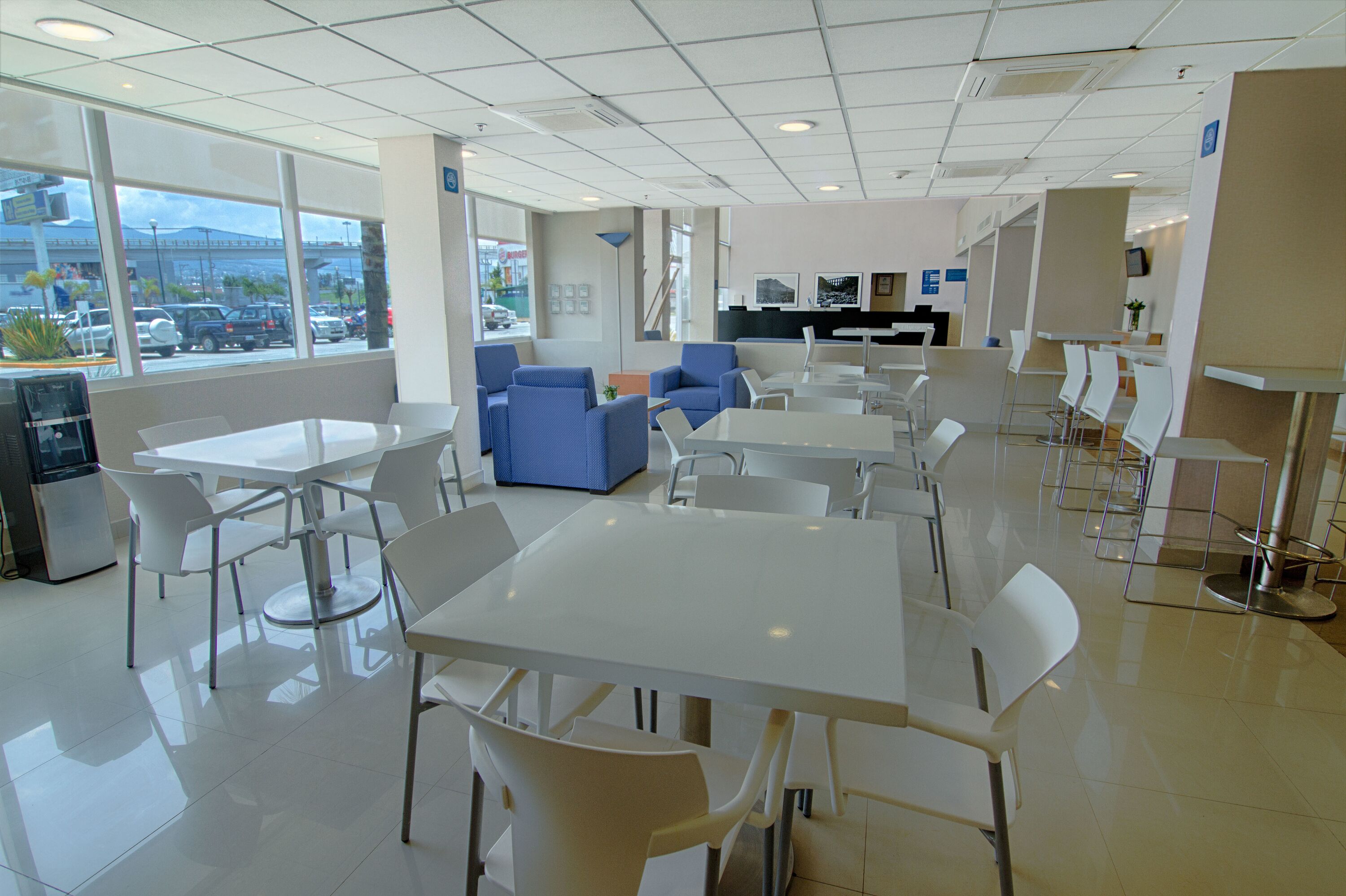 Un comedor moderno con mesas y sillas blancas, sofás azules y un amplio ventanal que ofrece vistas a coches aparcados y edificios.