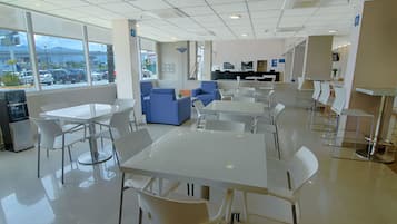 Un comedor moderno con mesas y sillas blancas, sofás azules y un amplio ventanal que ofrece vistas a coches aparcados y edificios.