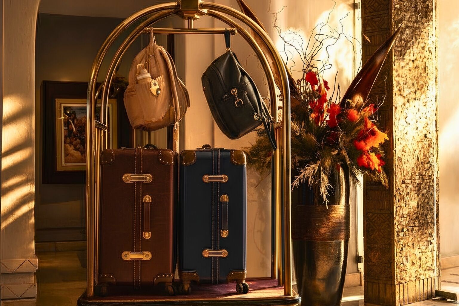 A vintage-style luggage rack with two suitcases and a handbag, a vase with red flowers, and a framed picture on the wall.