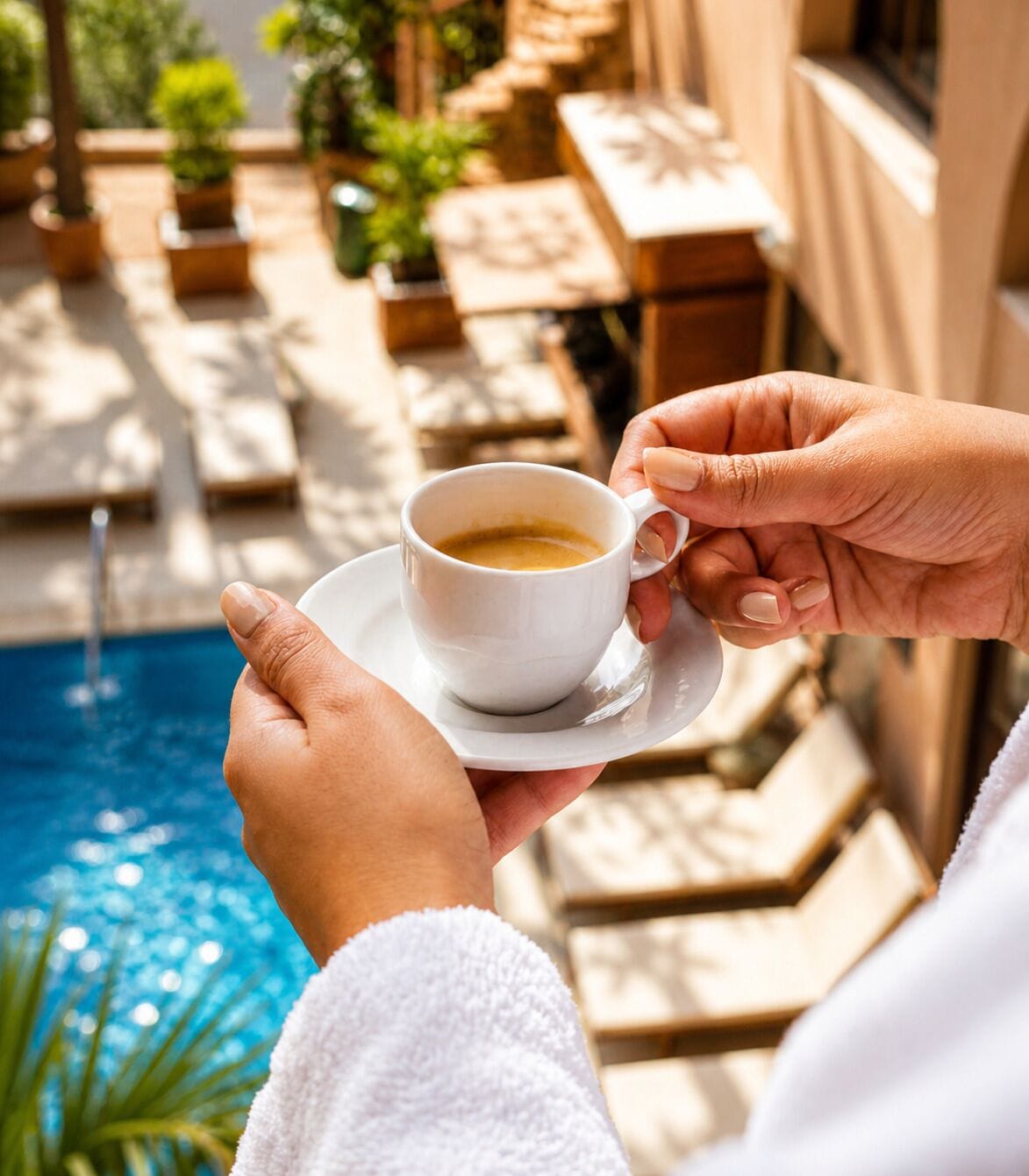 Una persona sostiene una taza blanca de café con platito, cerca de una piscina con fuente, rodeada de plantas en macetas y mobiliario exterior.