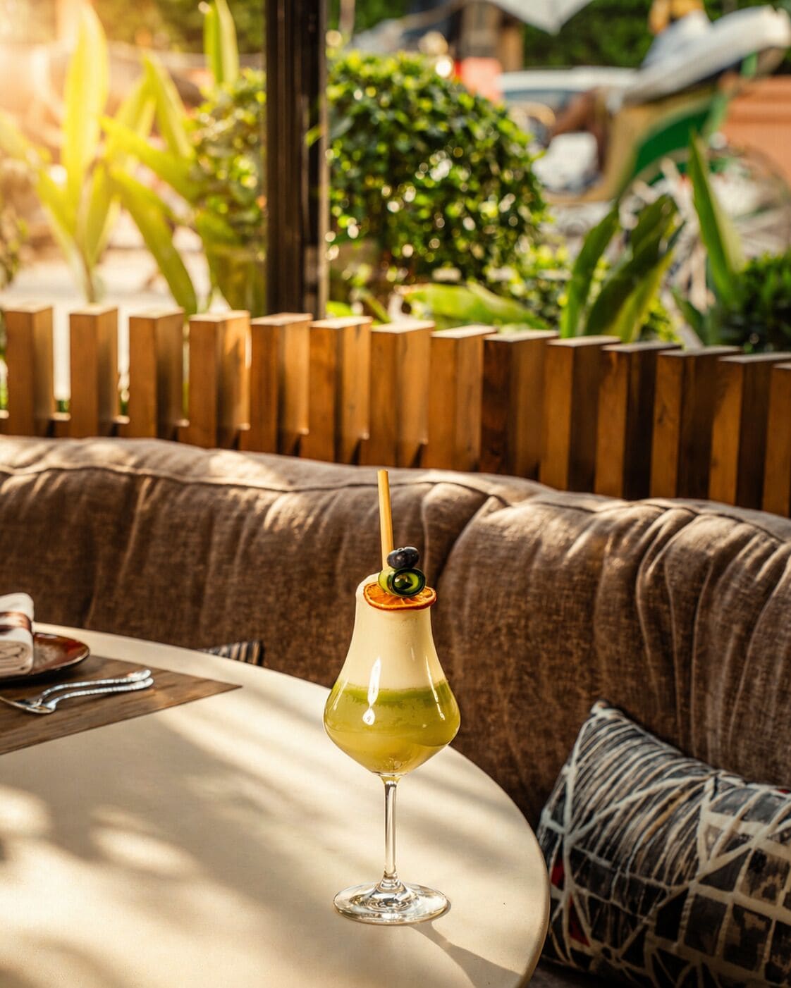 A cocktail in a glass with a straw, placed on a table in a seating area with a wooden fence and greenery in the background.