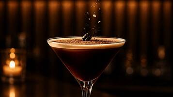 A martini glass with a dark liquid and a frothy top, placed on a surface with scattered coffee beans.