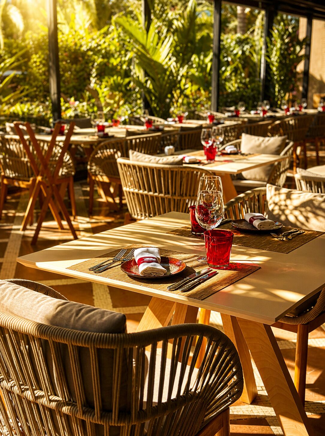 An outdoor dining area with wicker chairs and tables, some with red glasses and plates, and a tropical backdrop.