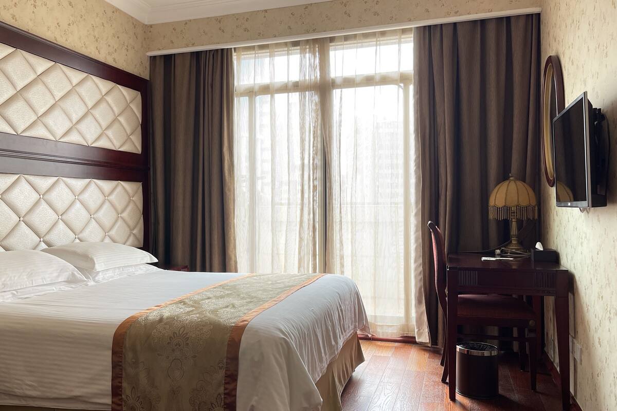 A hotel room with a large bed, a wooden headboard, a desk with a lamp, and a television on the wall.