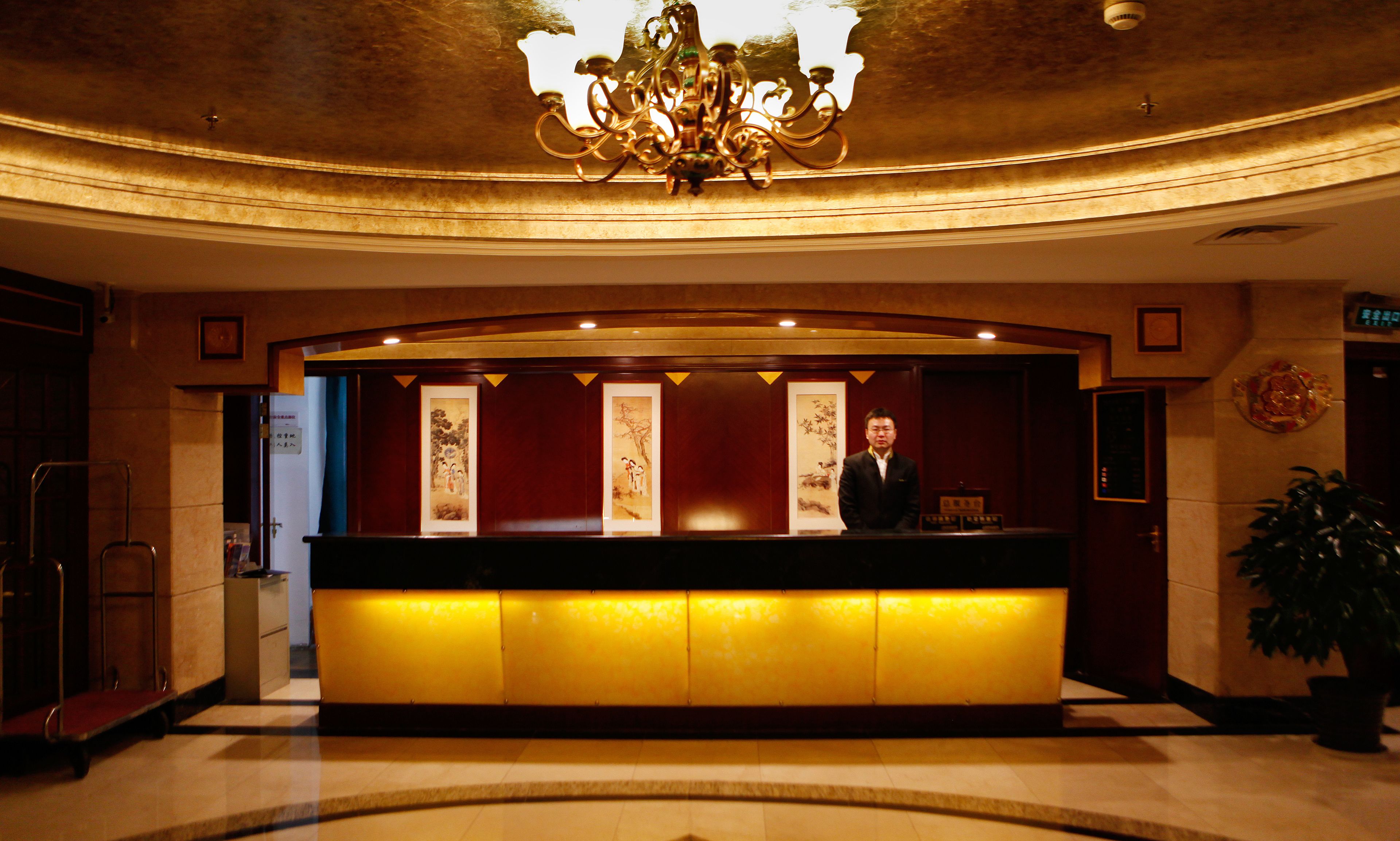 A hotel reception desk with a chandelier, framed artwork, and a potted plant.