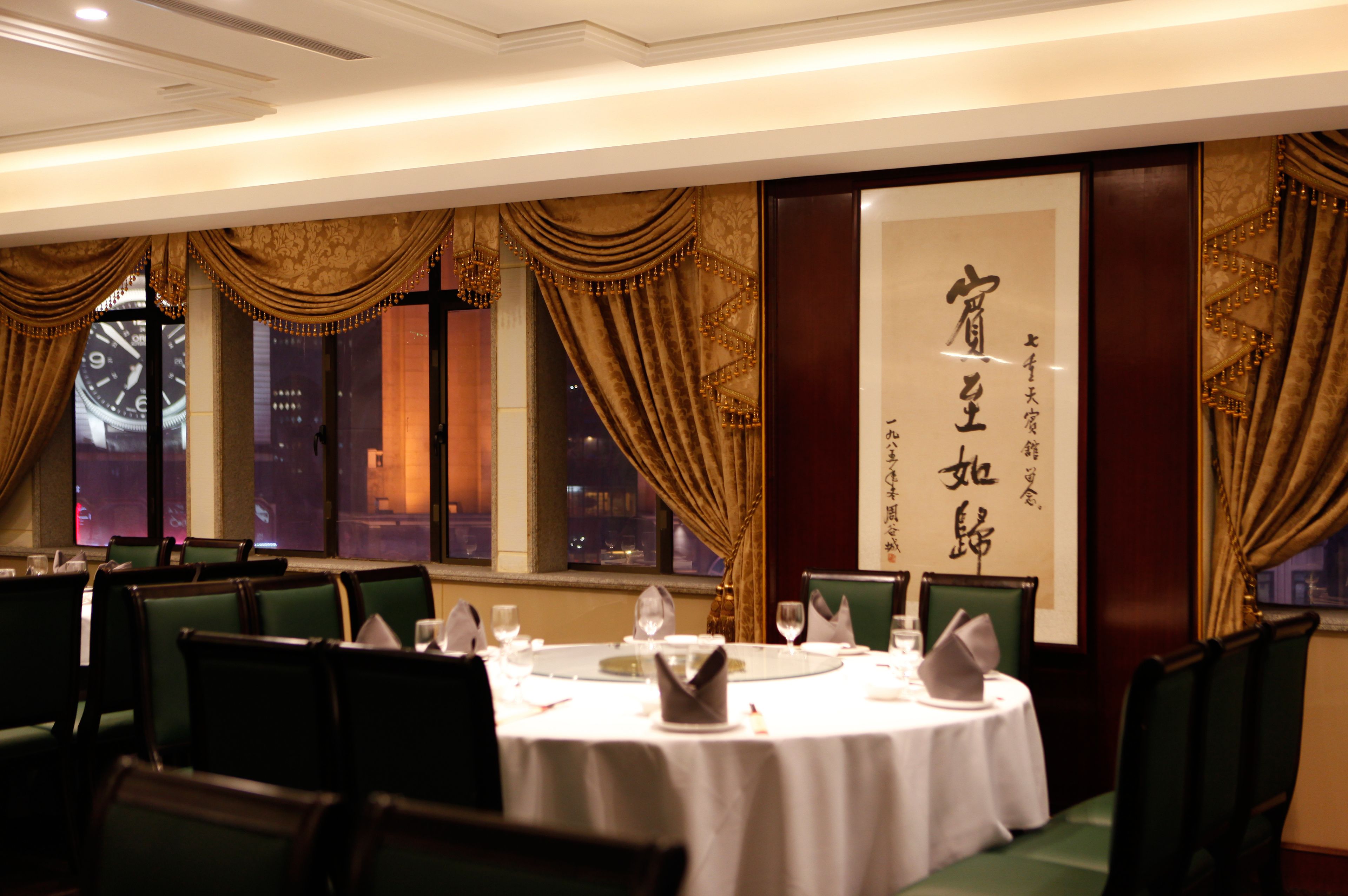 A dining area with tables set for service, large windows with curtains, and a wall with Chinese characters.