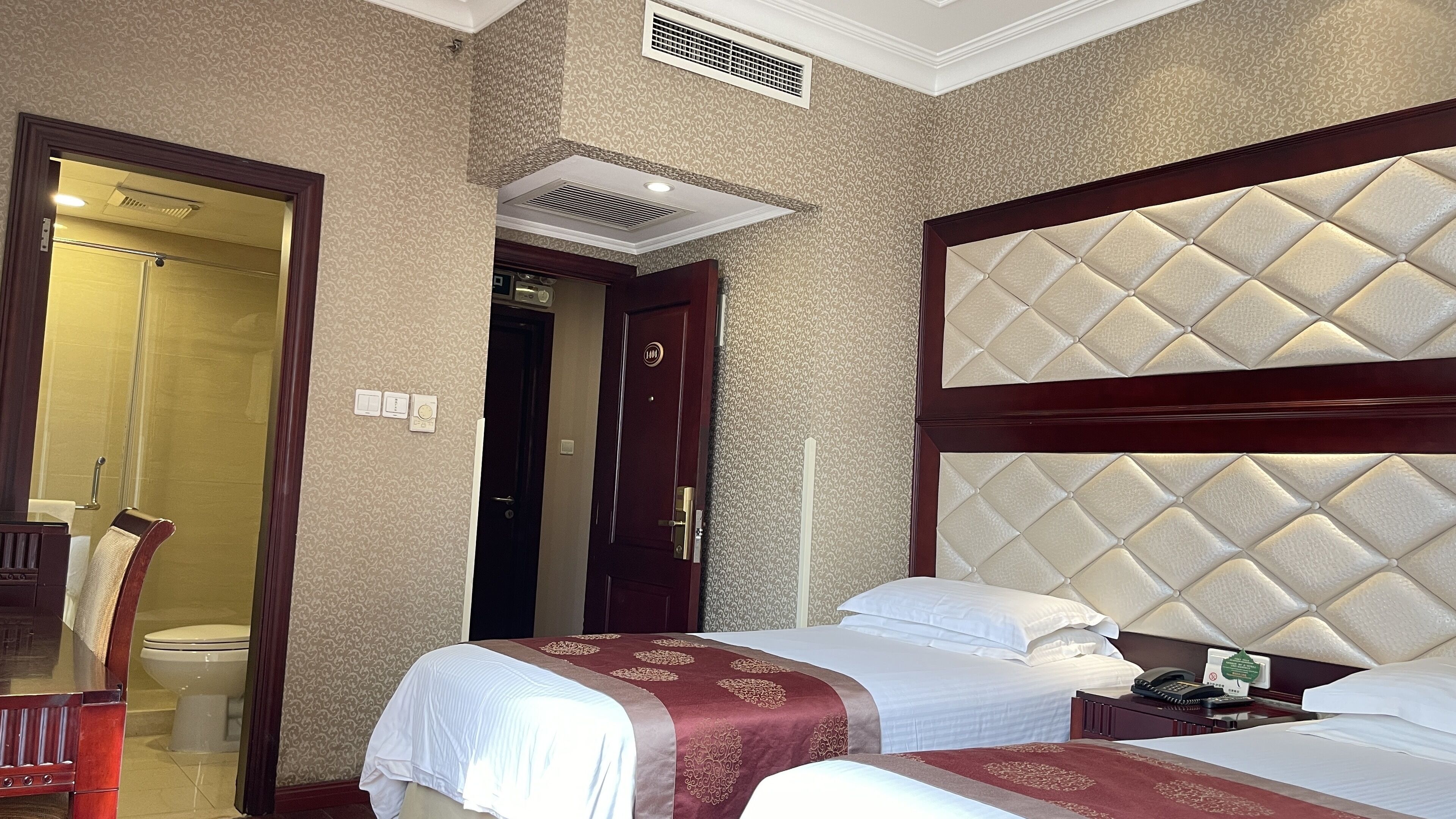 Habitación de hotel con dos camas, cabecero de madera, baño con inodoro y ducha, y una puerta que da a otra habitación.