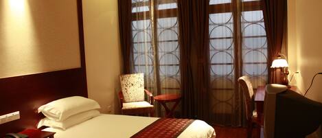 Quarto de hotel com uma cama, duas cadeiras, uma mesinha, escrivaninha com luminária e janela com cortinas.