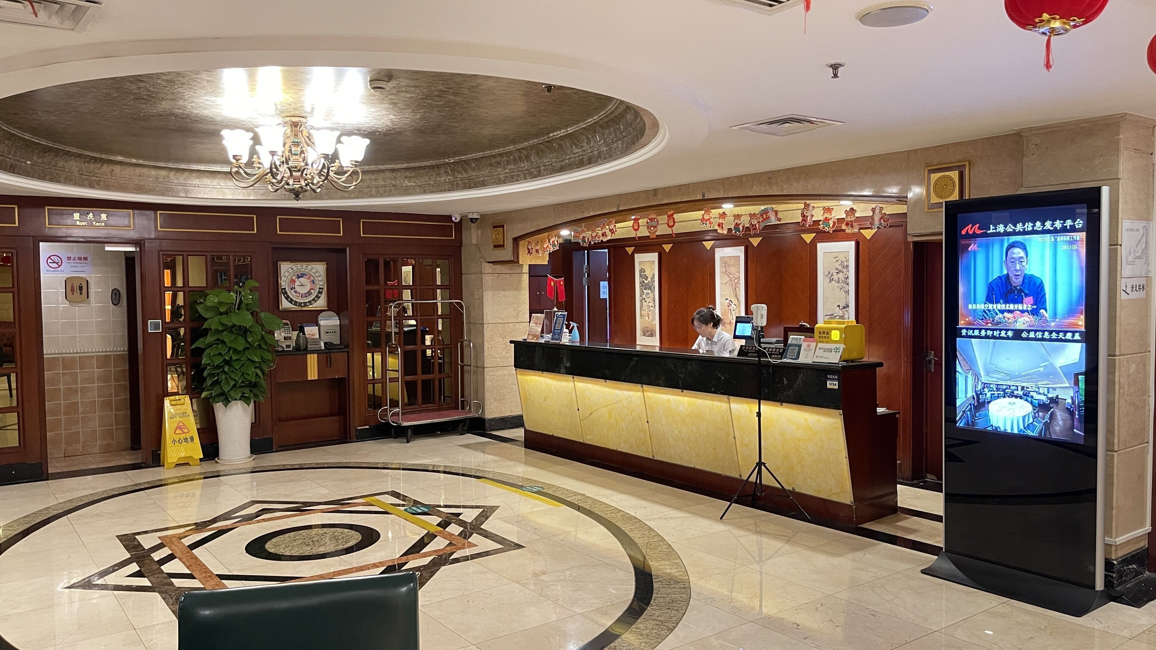 Zona de recepción de hotel con piso de mármol, mostrador de recepción, pantalla de televisión y candelabro.