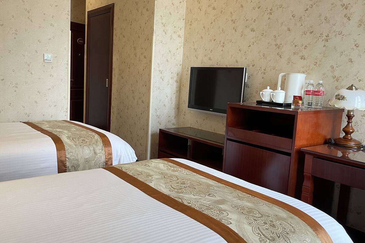 A hotel room with two beds, a TV, a desk with a lamp, and a shelf with toiletries.