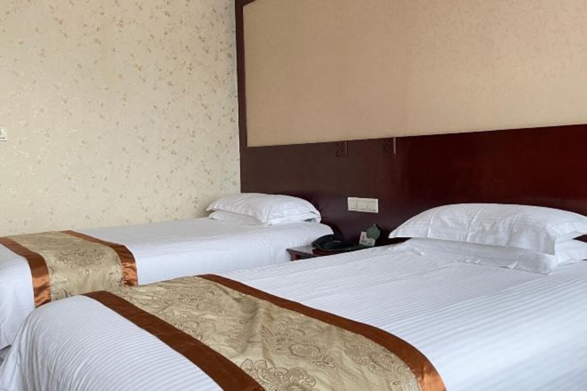 Habitación de hotel con dos camas, cada una con ropa blanca y un borde decorativo marrón. Entre las camas hay una mesita de noche con una lámpara y un teléfono. La habitación tiene un cabecero de madera y un papel tapiz estampado en la pared.