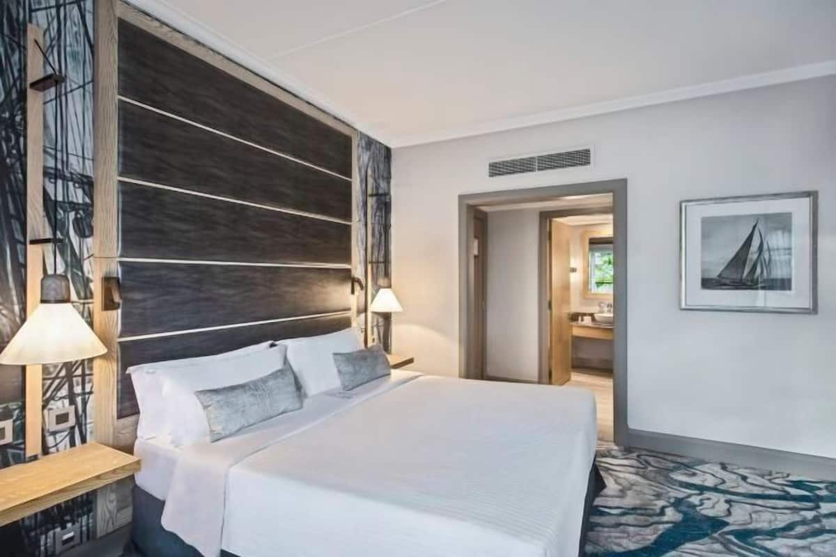 Una habitación de hotel moderna con una cama grande, lámparas de noche, un cabecero de madera, un cuadro enmarcado en la pared y una alfombra estampada.