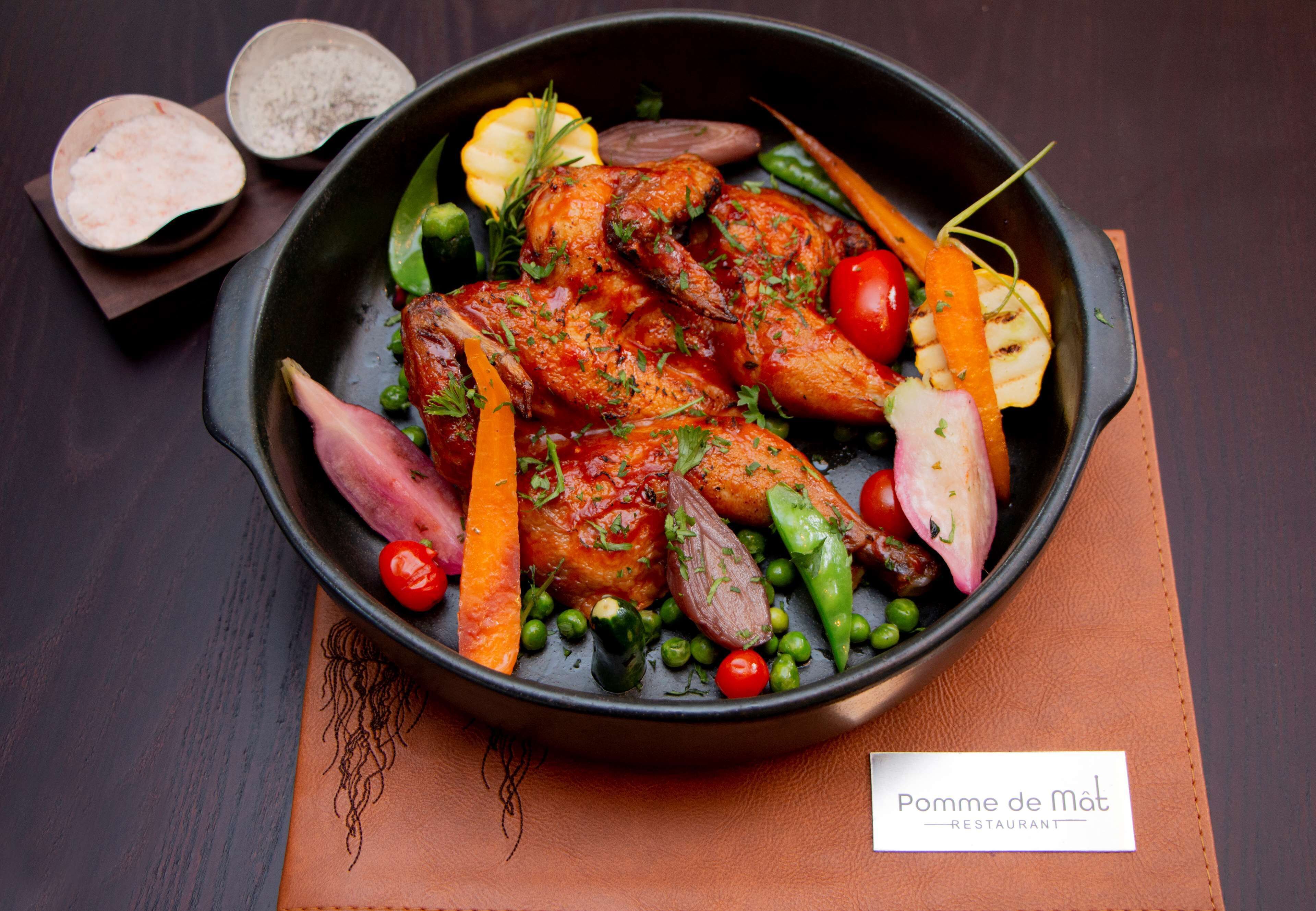 Un plato de pollo asado con verduras en una sartén negra, sobre una mesa marrón con una tarjeta de identificación que dice 
