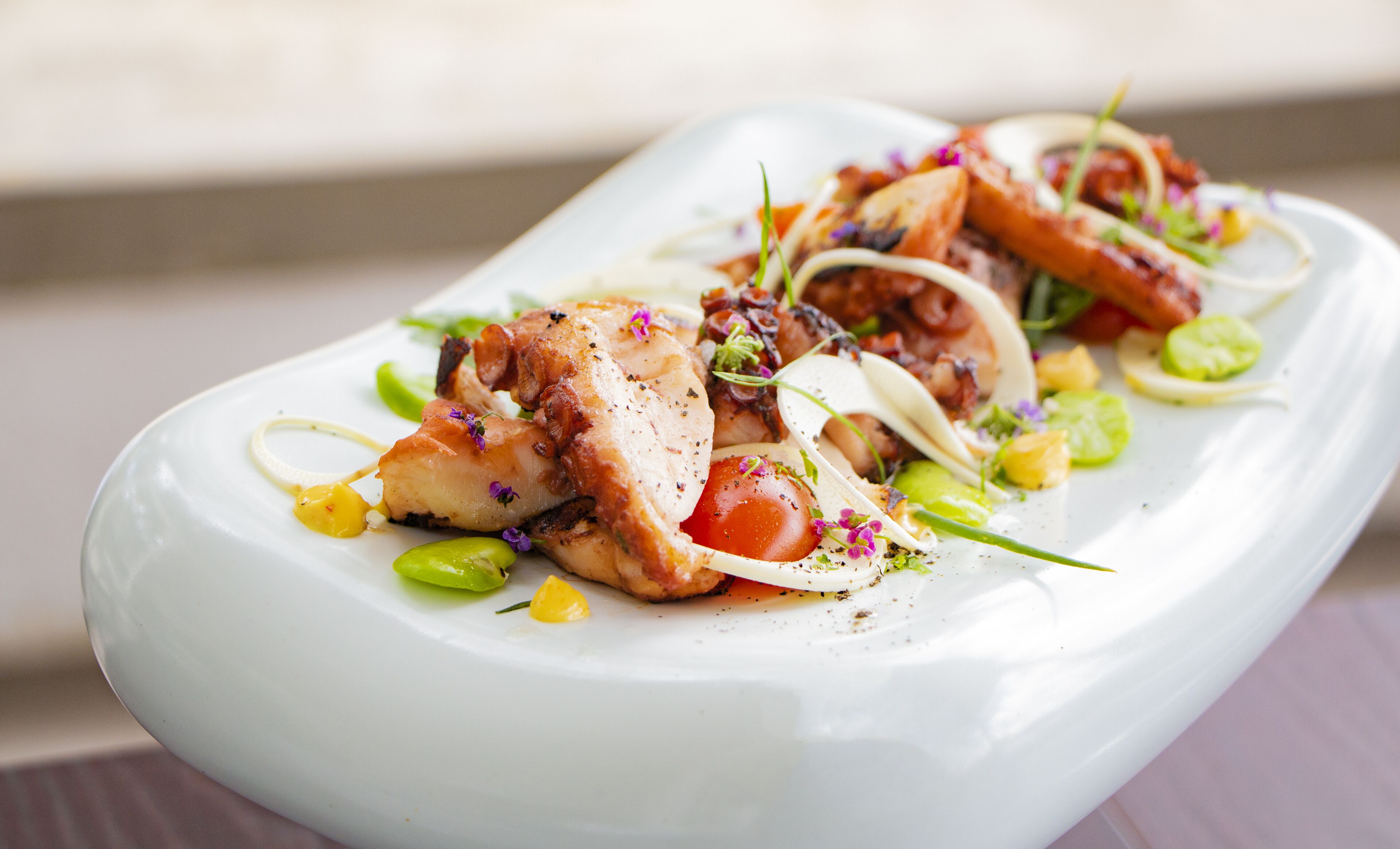 A white rectangular plate with a seafood dish, garnished with green leaves and purple flowers.